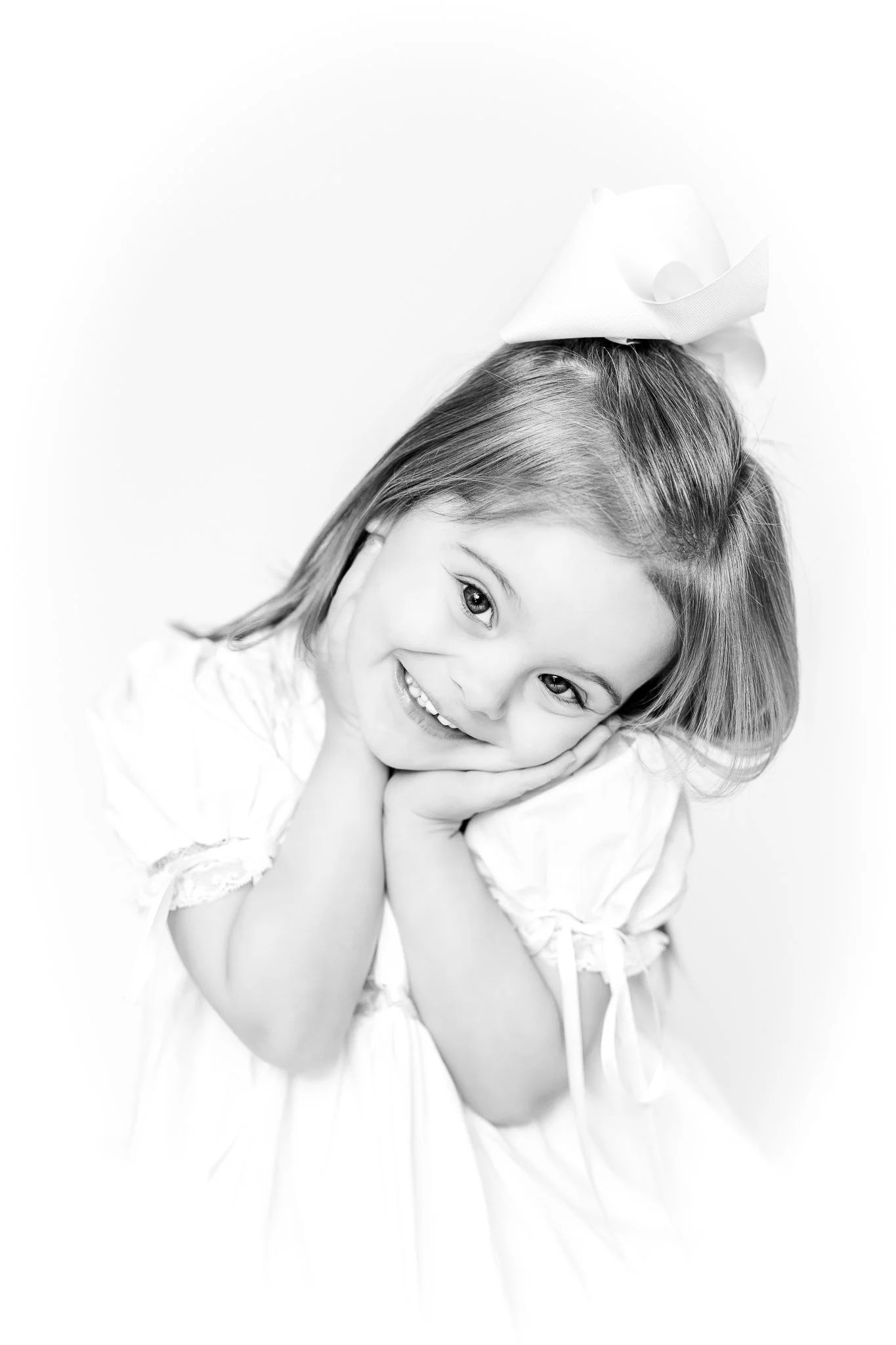 Black and white photo of a smiling young girl with a big hair bow, resting her chin in her hands.