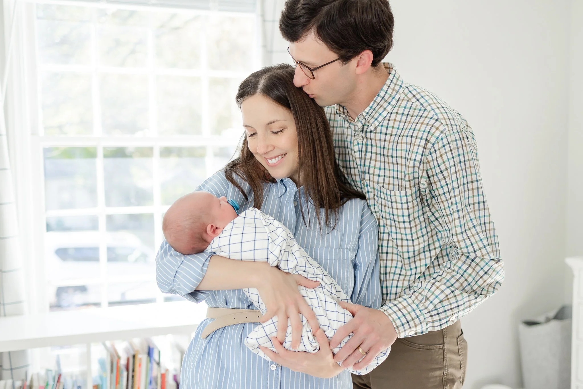 A couple lovingly holding their newborn baby wrapped in a checkered blanket, standing in a sunlit room.
