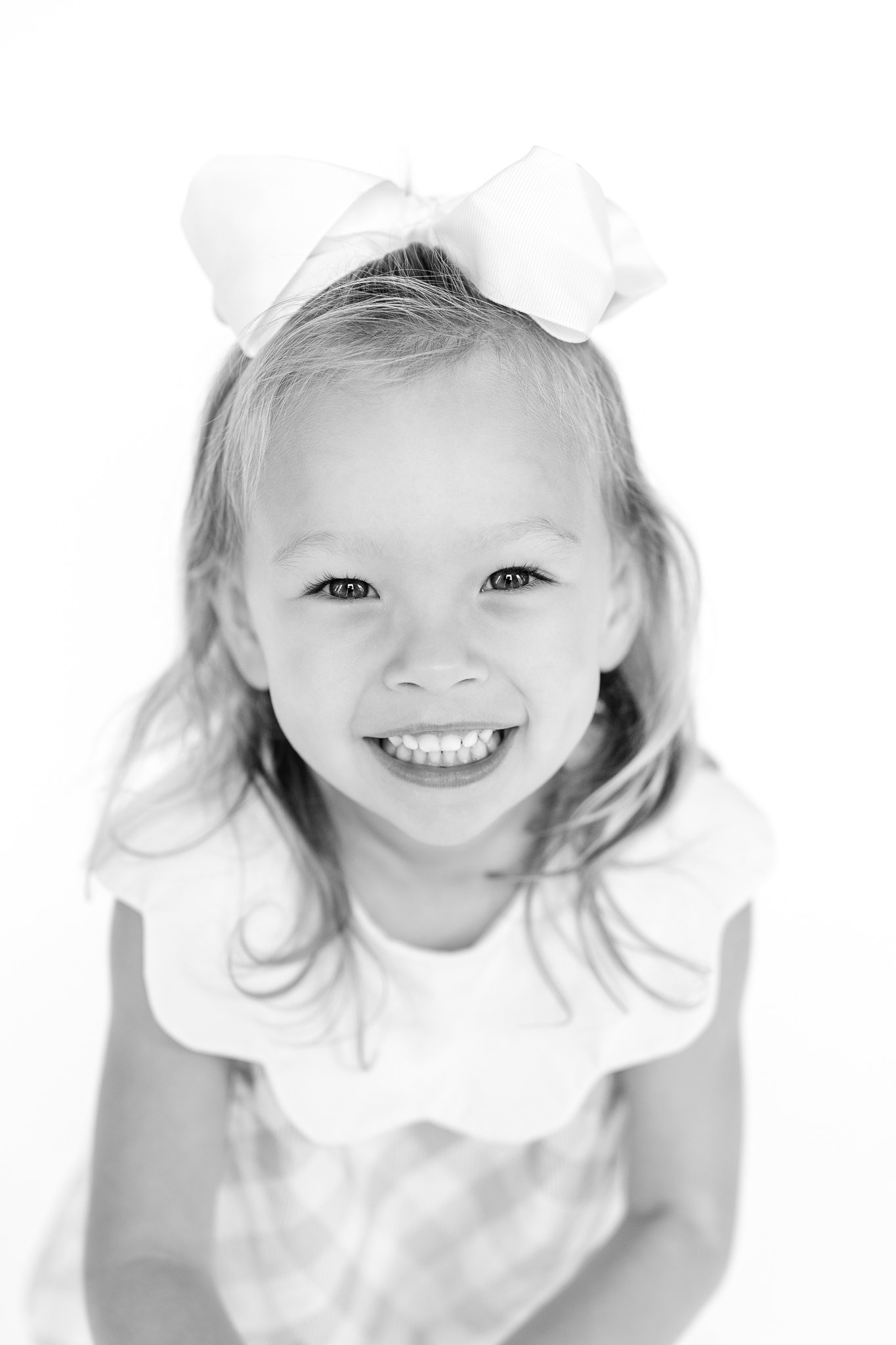 Black and white photo of a smiling child with long hair and a large bow, wearing a dress.