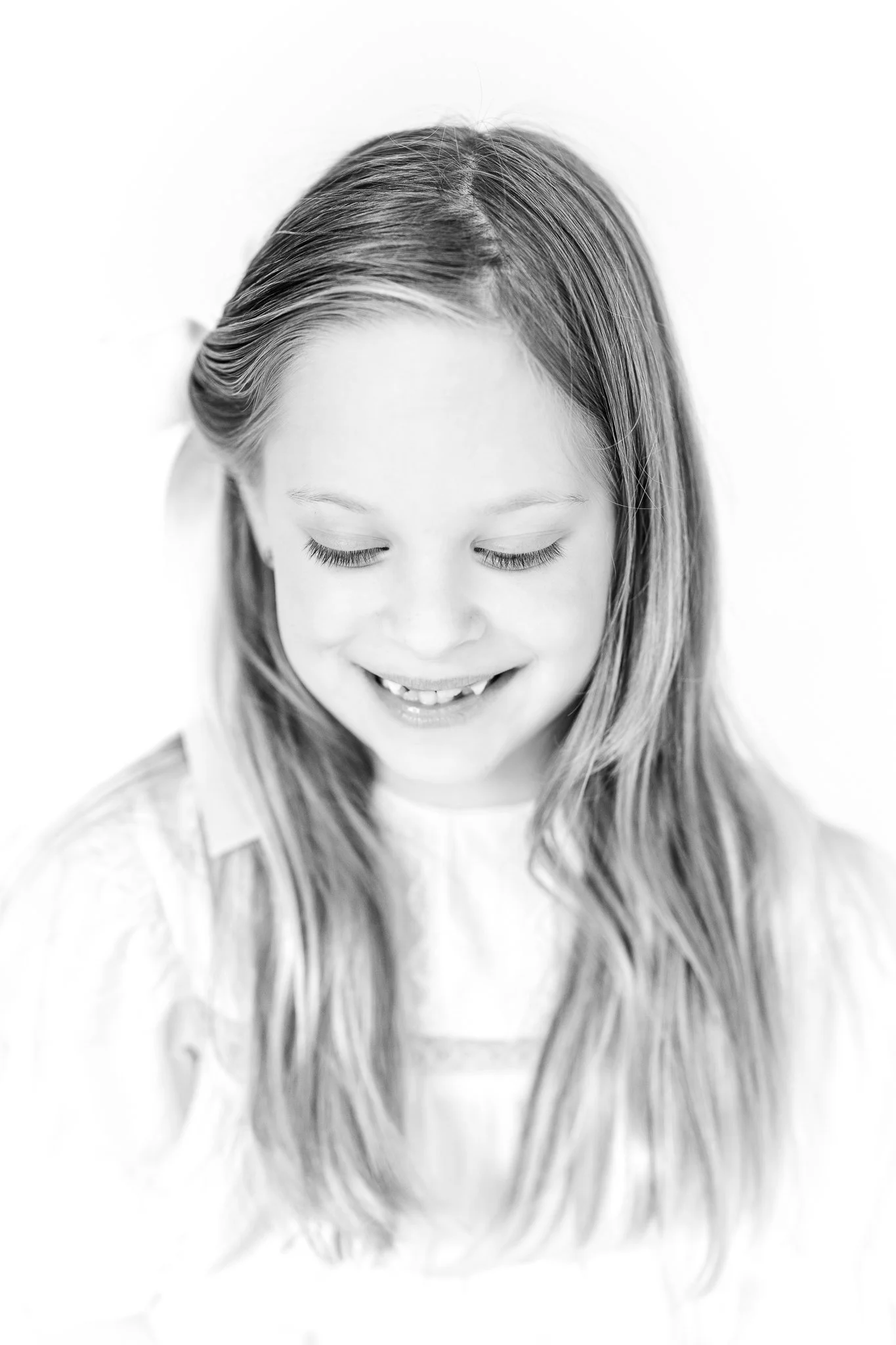 Black and white portrait of a young girl smiling with her eyes closed, wearing a light-colored dress and a ribbon in her hair.