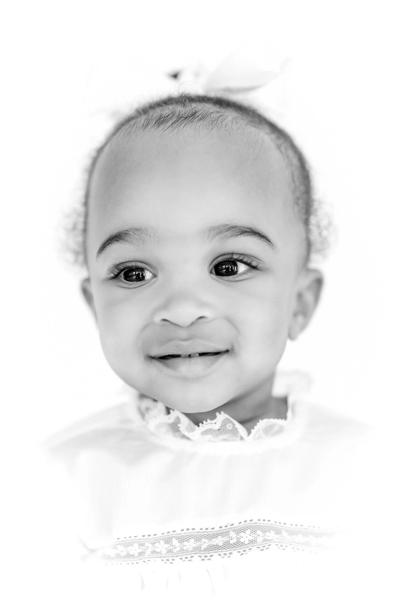 Black and white portrait of a smiling toddler wearing a lace-trimmed shirt with a bow in their hair.
