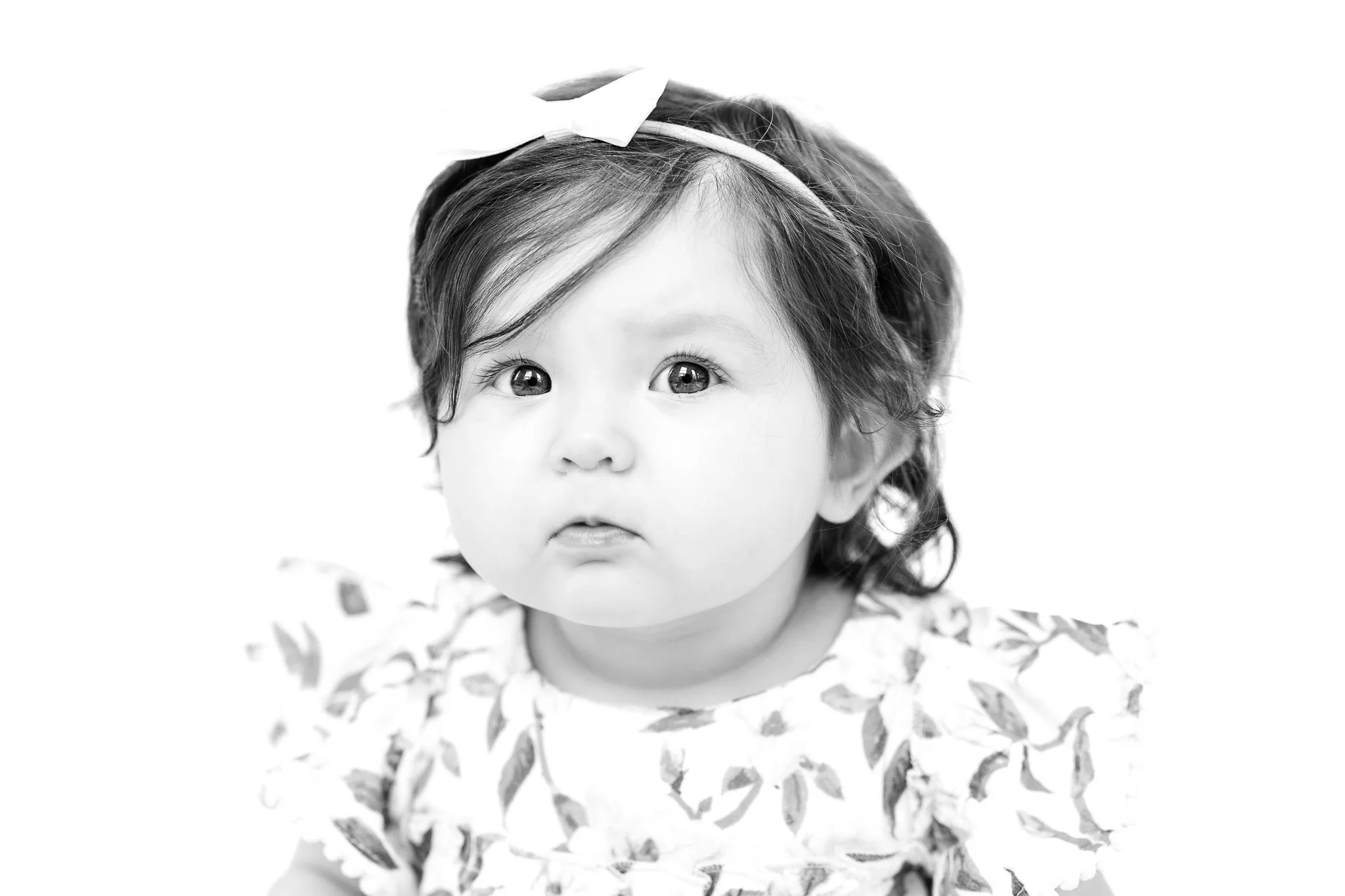 Black and white photo of a baby with a headband and patterned outfit, looking directly at the camera.
