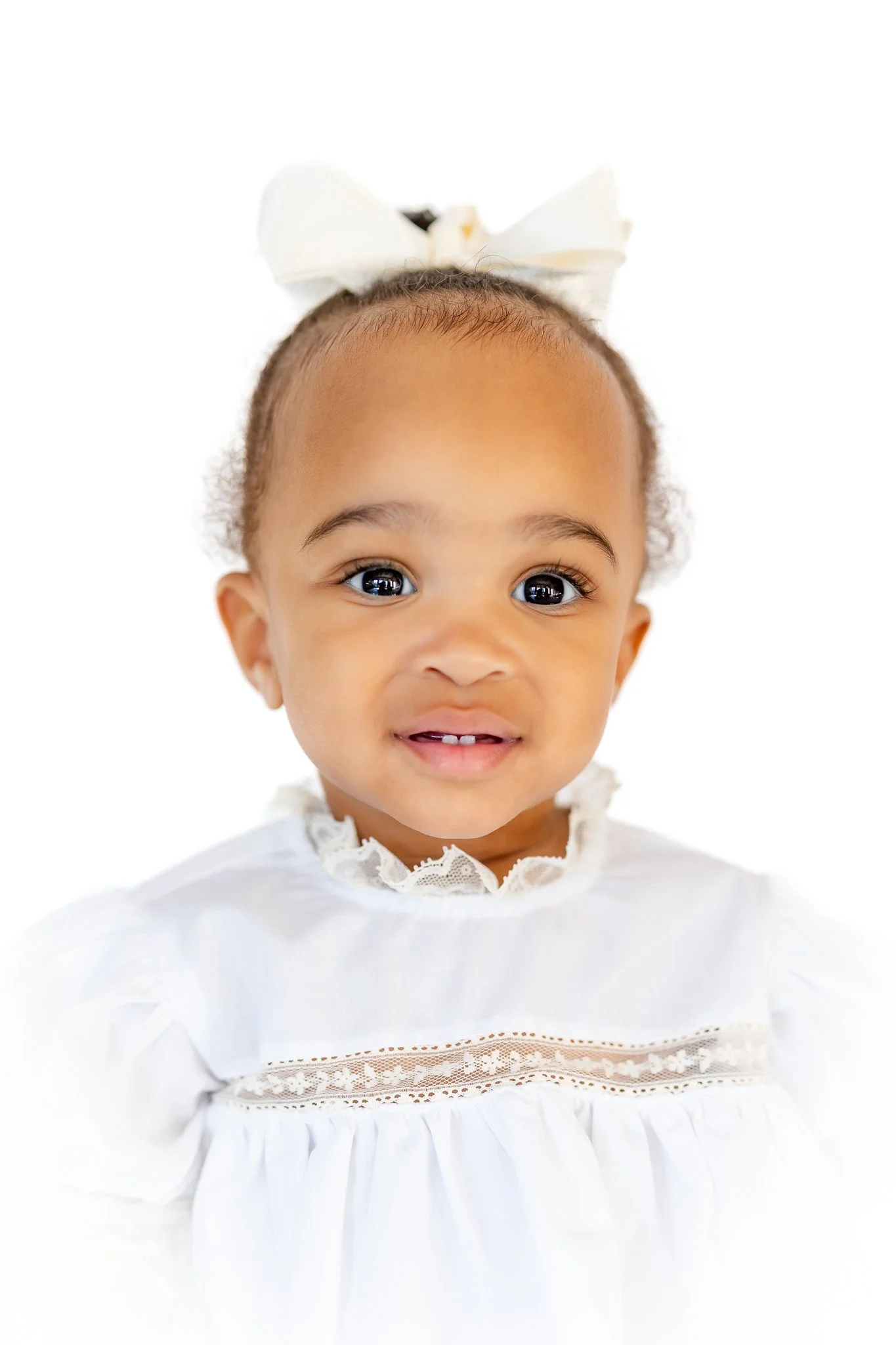 Smiling toddler wearing a white lace dress and a hair bow against a white background.