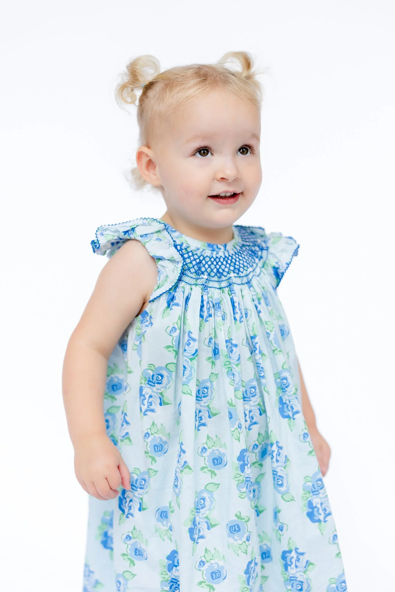 Young child with blonde hair in pigtails wearing a floral dress with blue flowers, smiling against a white background.