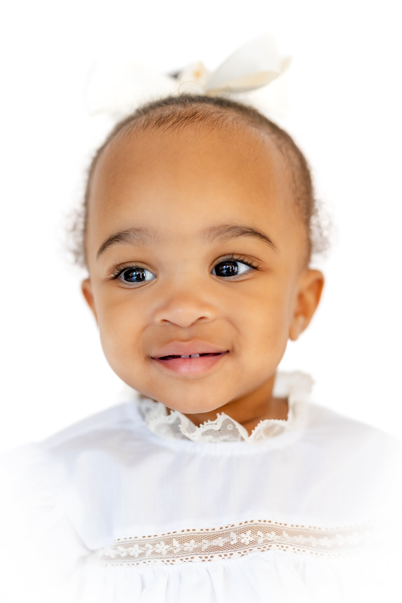 Smiling baby with a white dress and white bow.