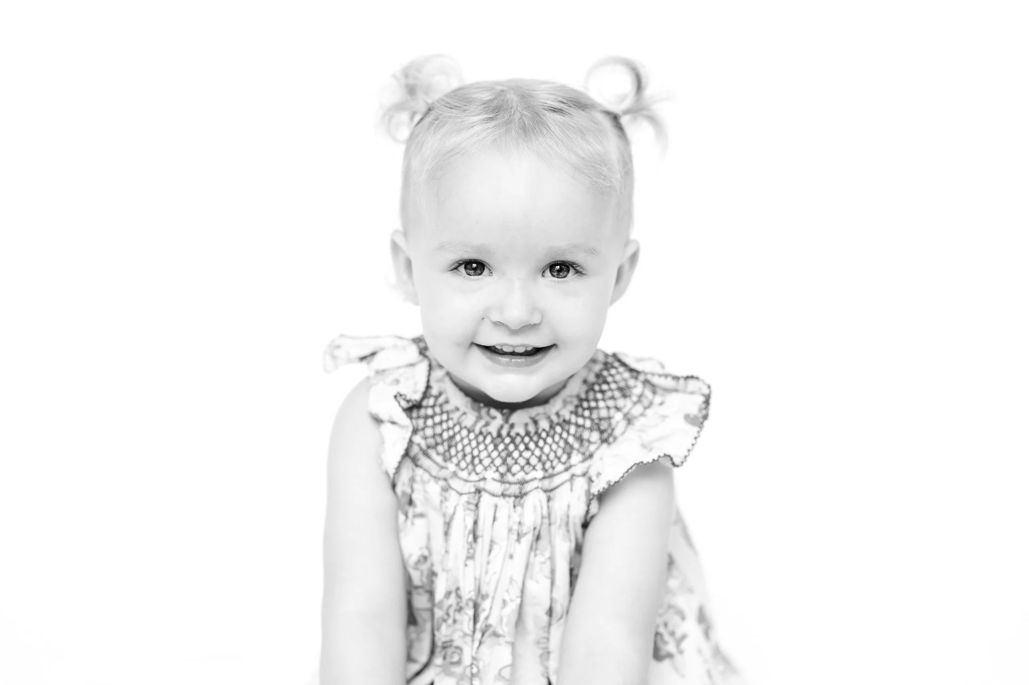 Black and white portrait of a smiling toddler with pigtails and a ruffled dress.