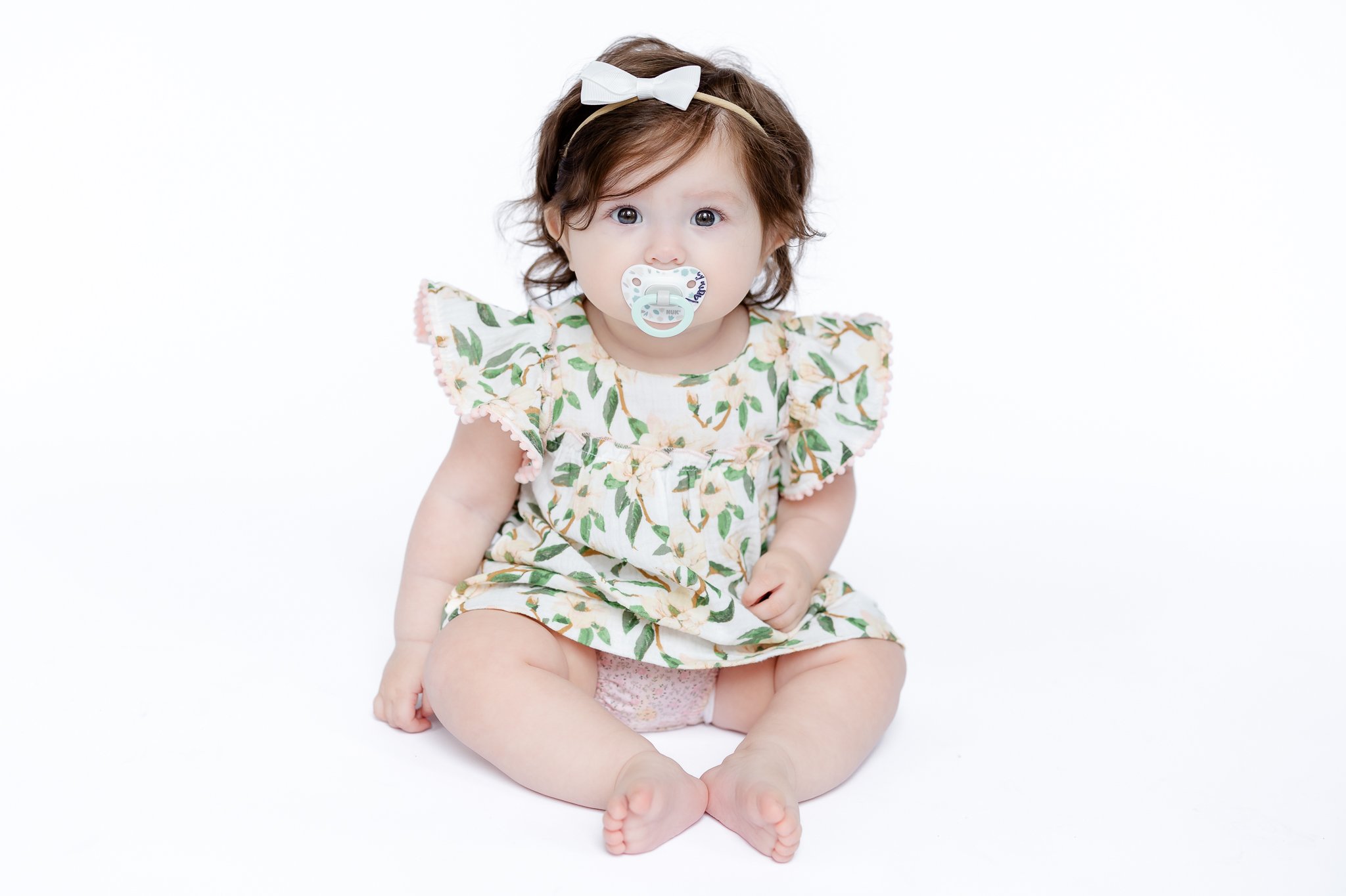 Baby wearing floral dress and pacifier, sitting on white background.
