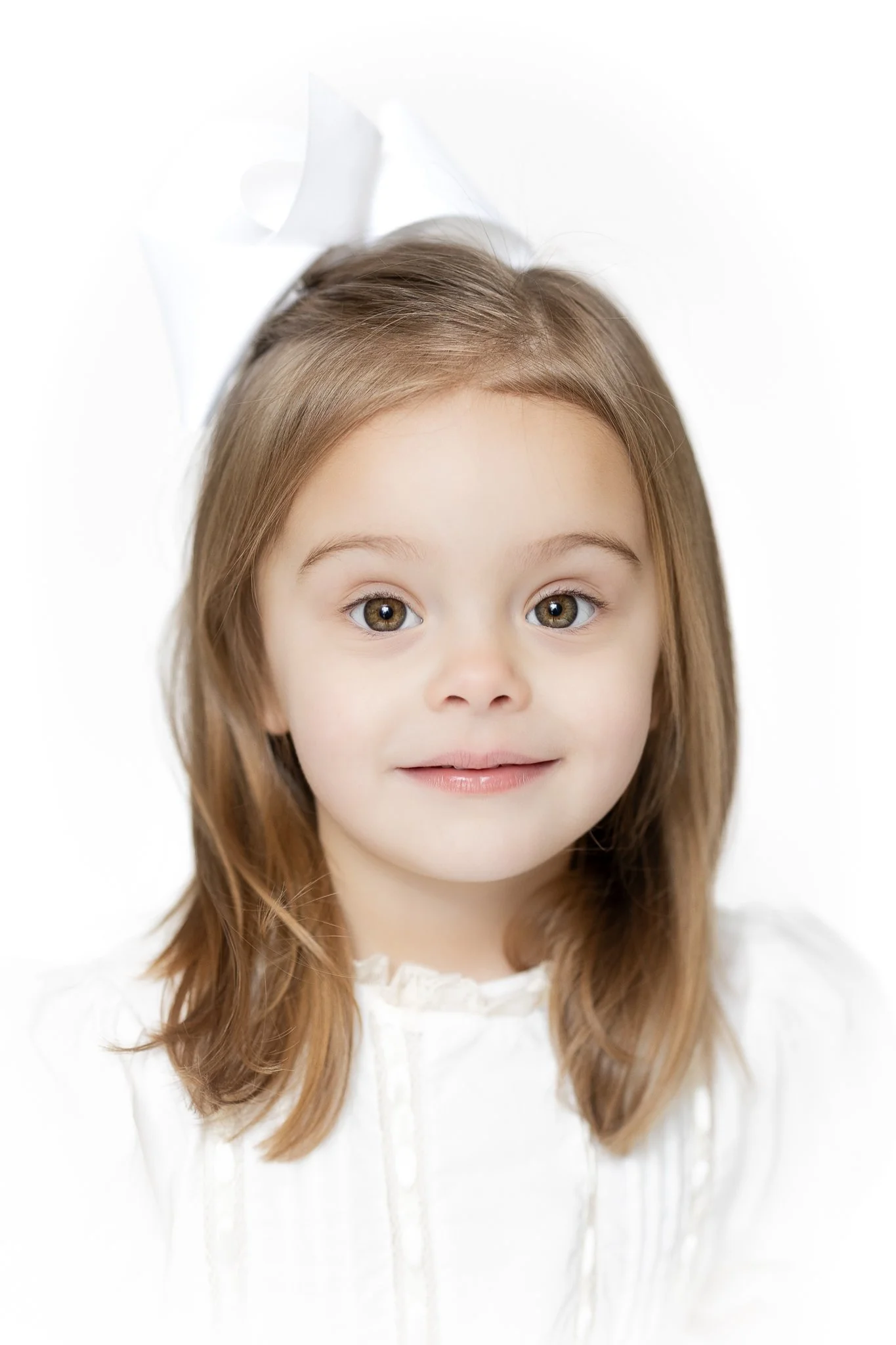 Young girl with long hair and a white bow, wearing a white dress, smiling slightly against a light background.