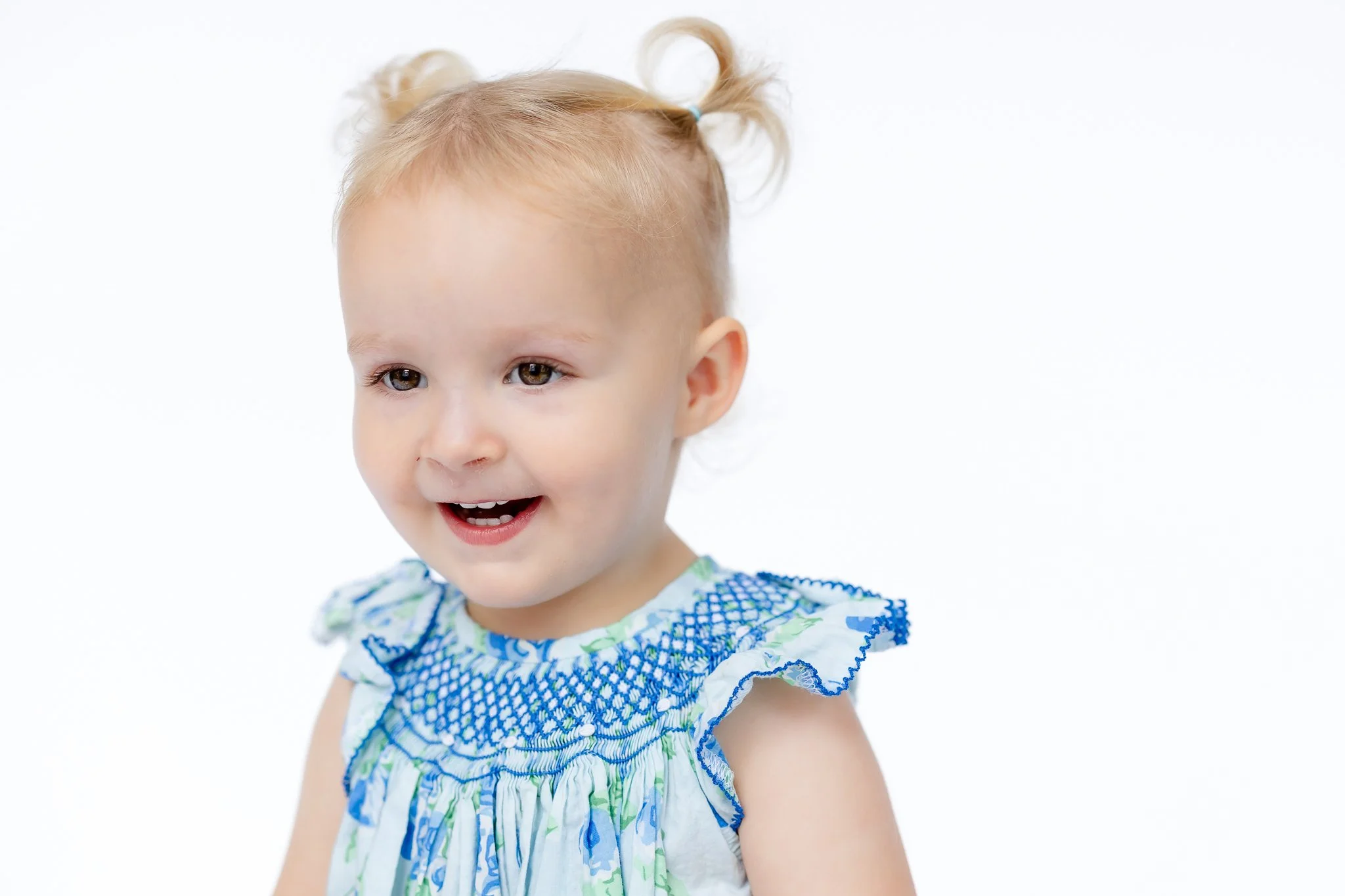 Smiling toddler with blonde hair in pigtails wearing a blue and white dress.