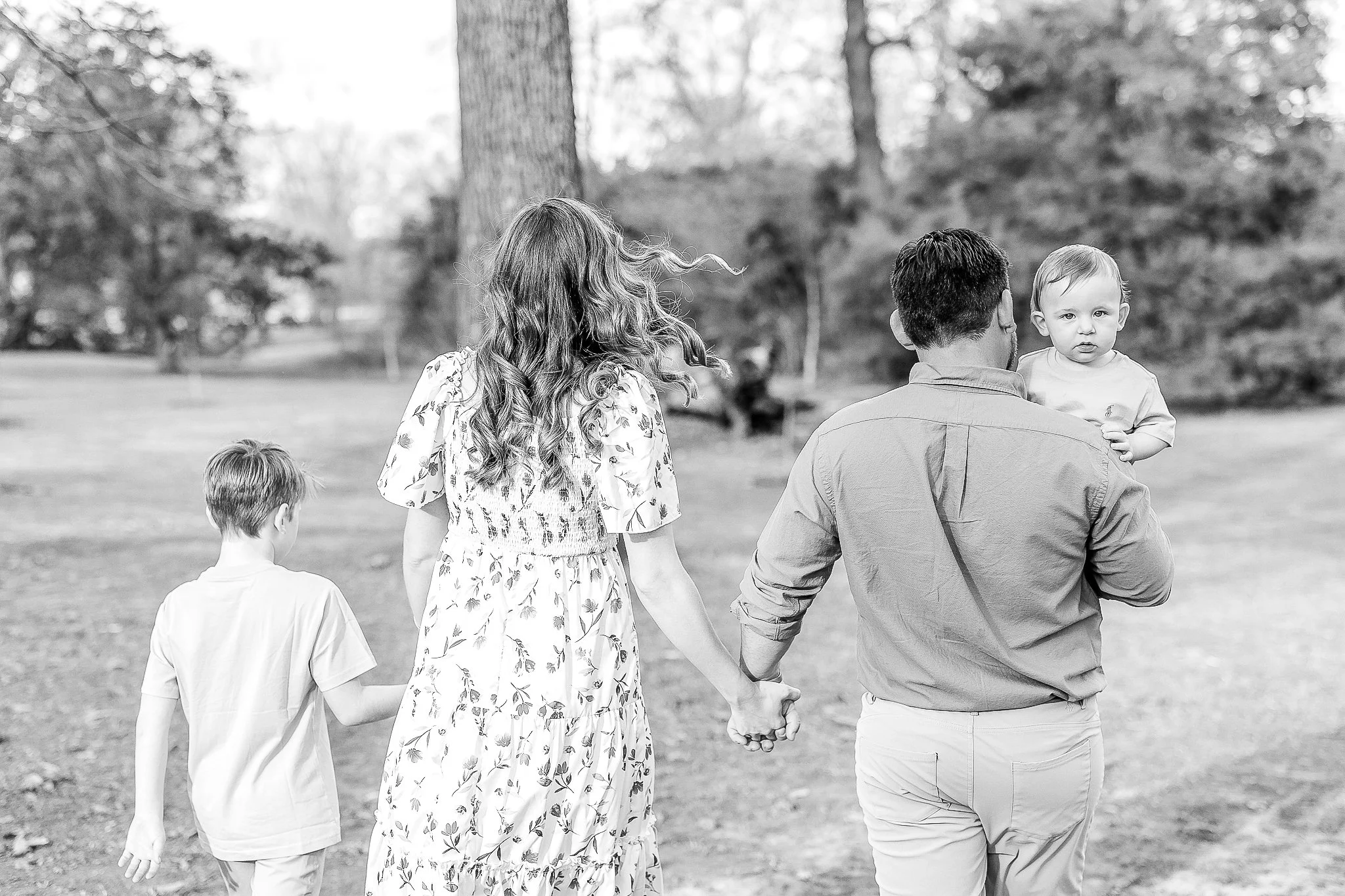 Black and white photo of a family walking in a park, with a woman in a floral dress holding hands with a man carrying a baby.
