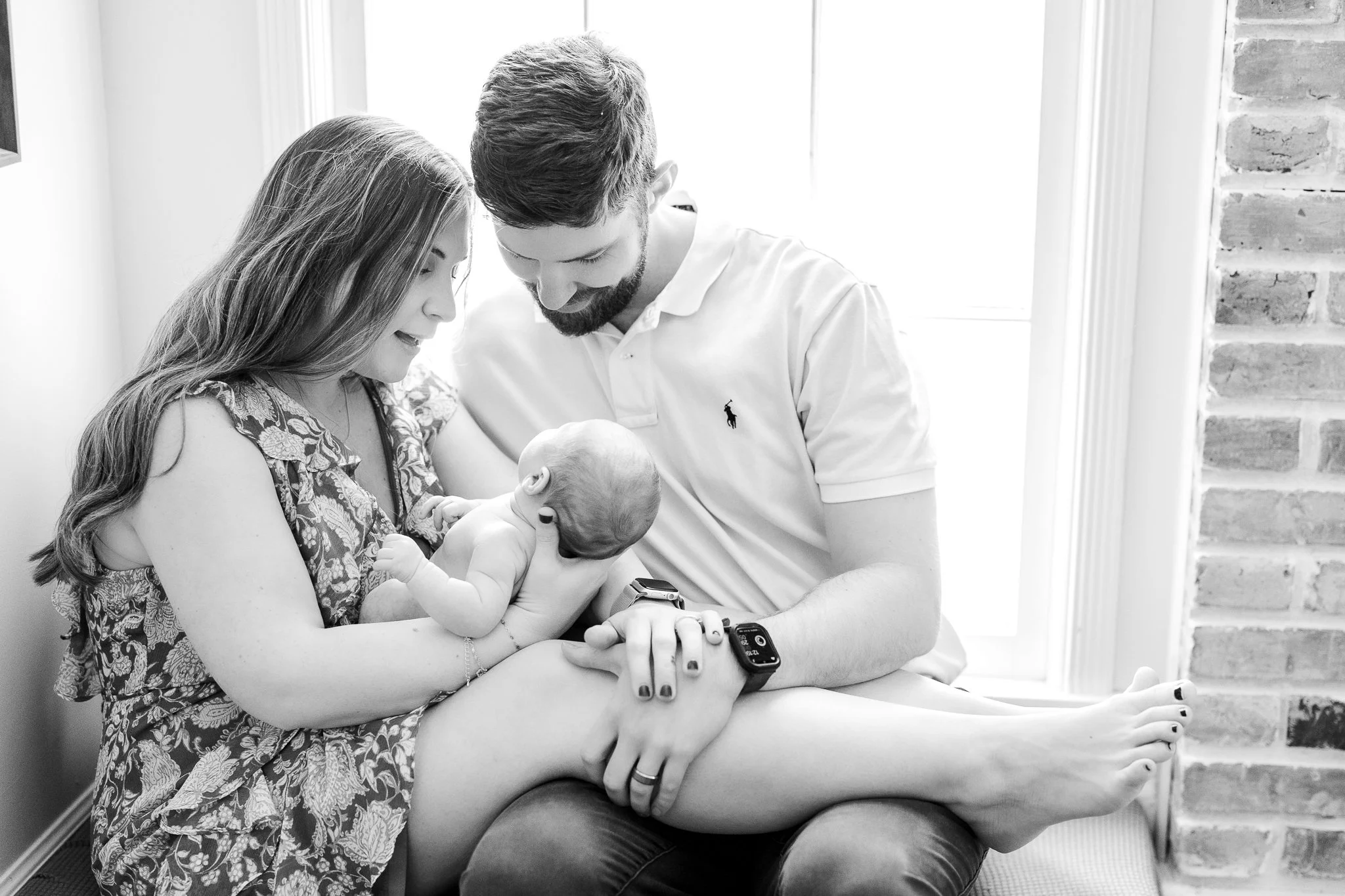 A black and white photo of a couple seated by a window, looking lovingly at their newborn baby, who is held by the mother.