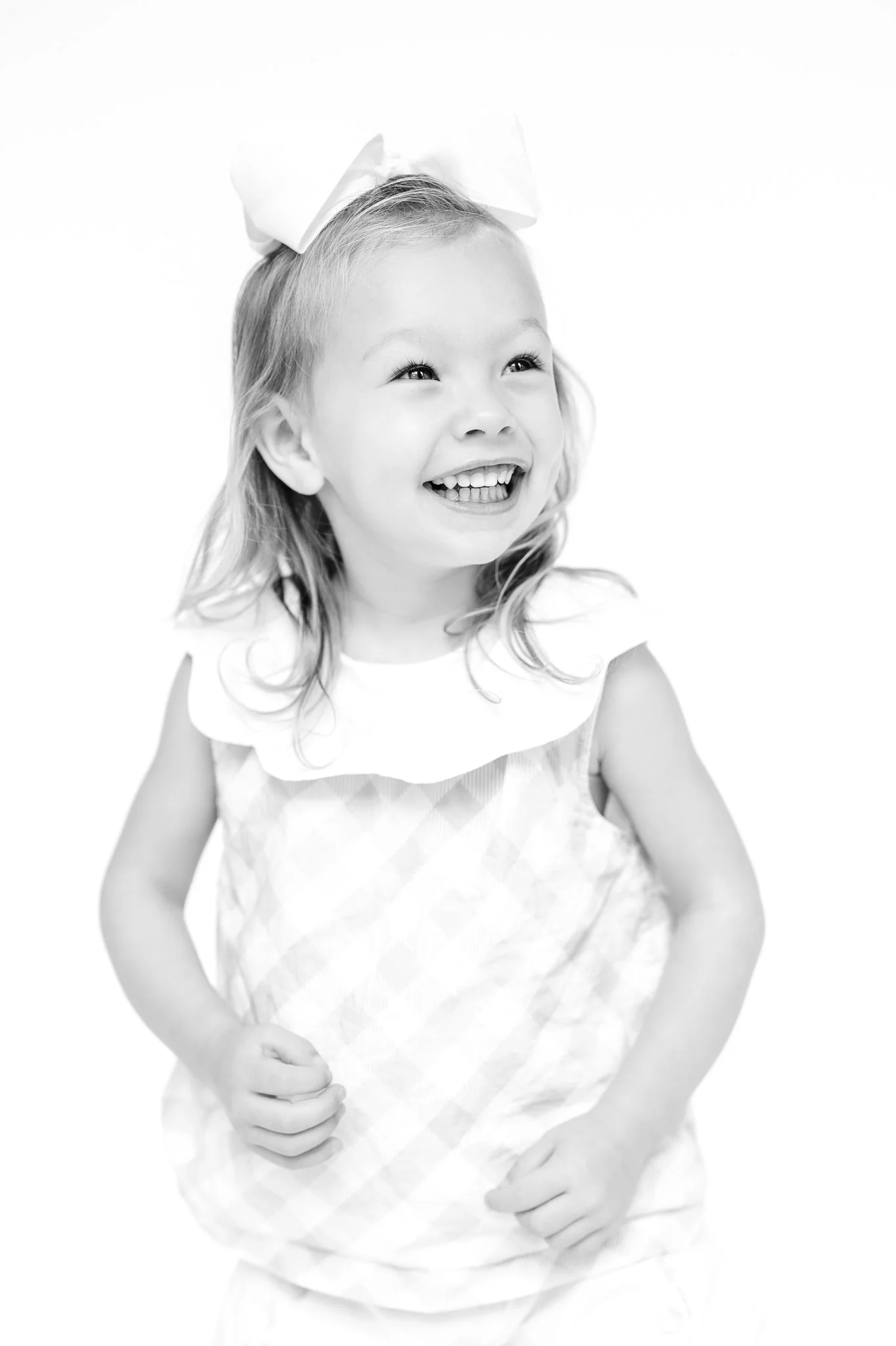 Black and white photo of a smiling young girl with a large bow in her hair, wearing a sleeveless top.