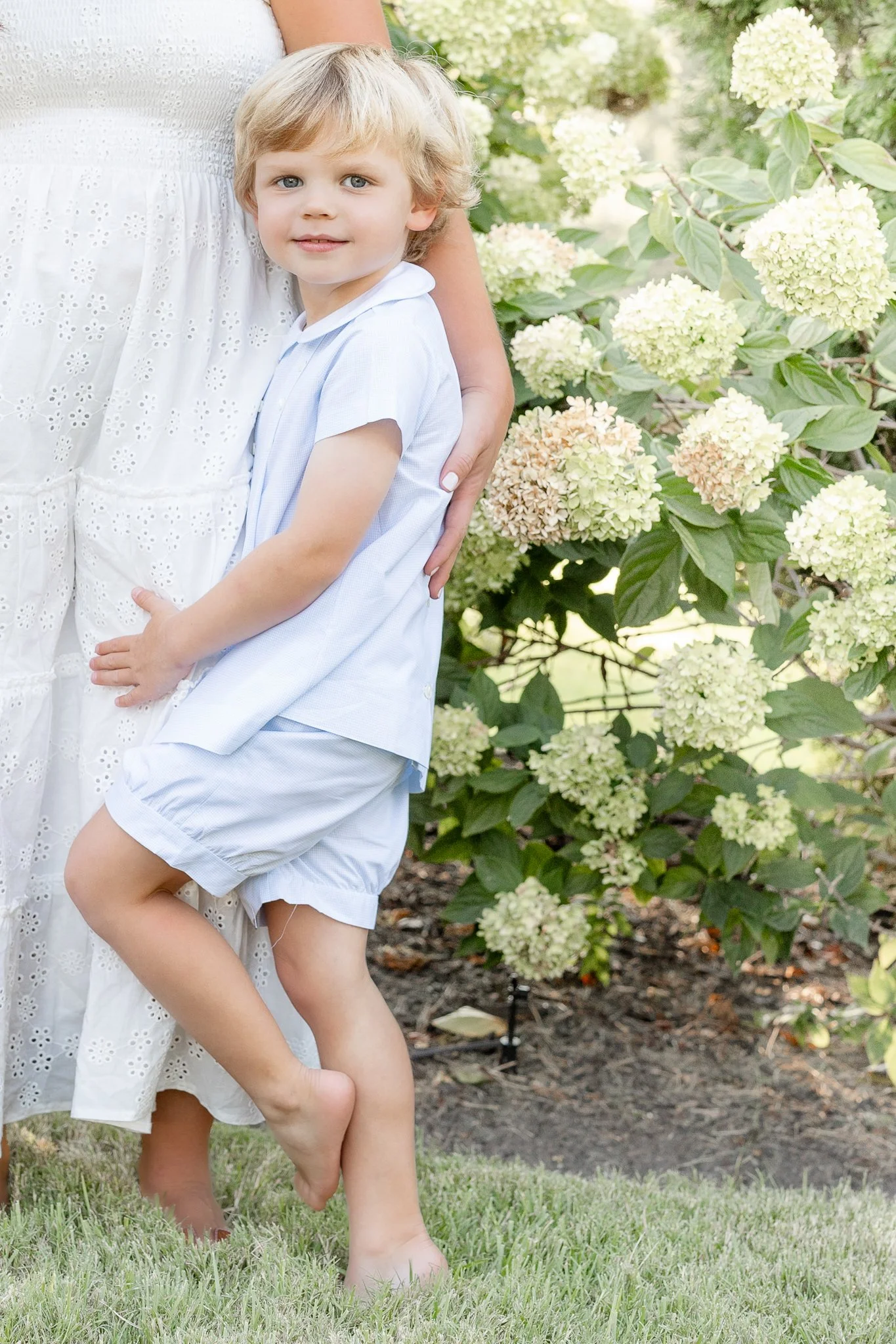 Child in light blue outfit standing barefoot on grass next to a person in a white dress, with flowering bushes in the background.