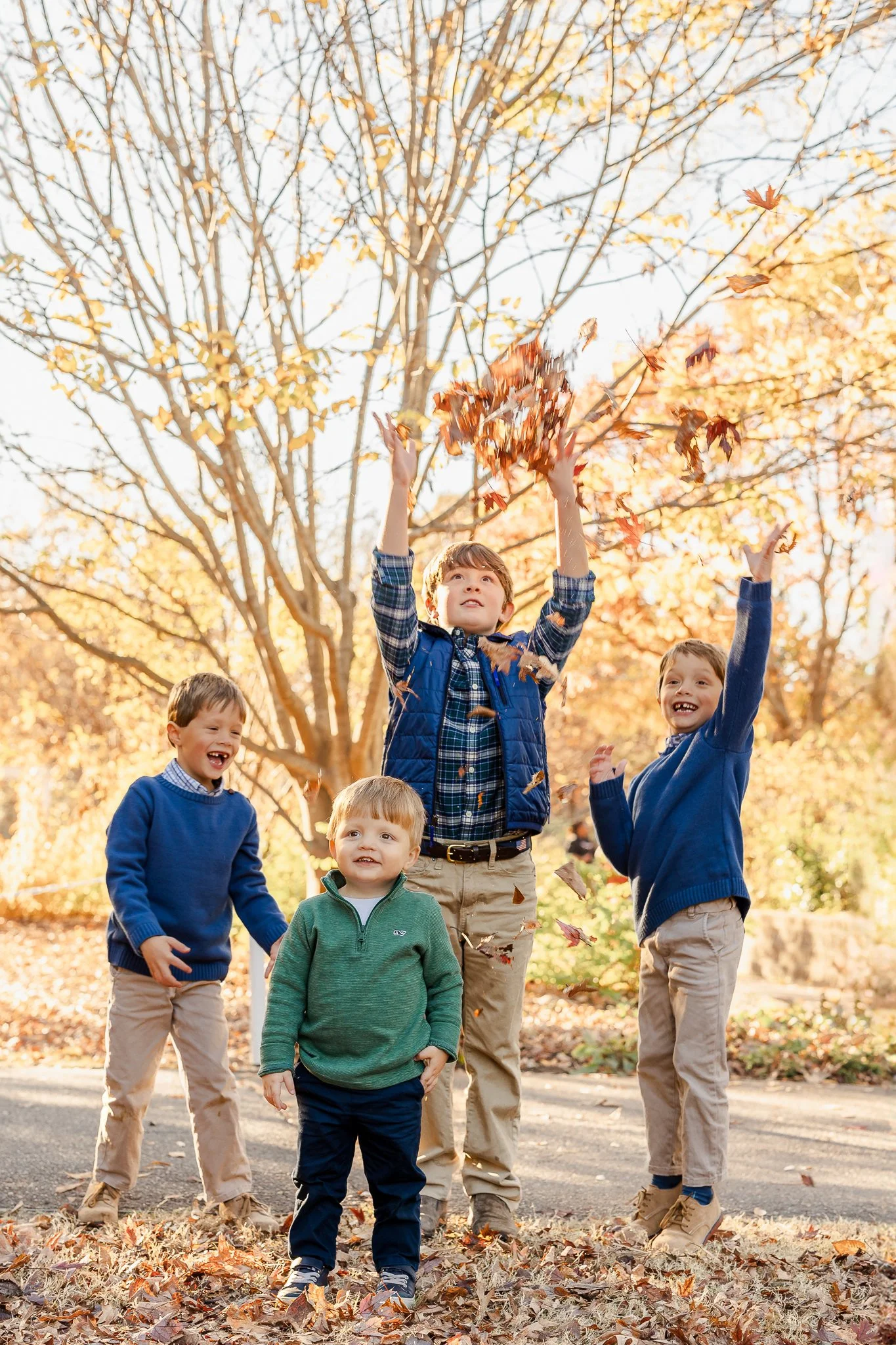 Four young boys playing with autumn leaves in a park, wearing casual fall clothing. Trees with yellow leaves in the background.