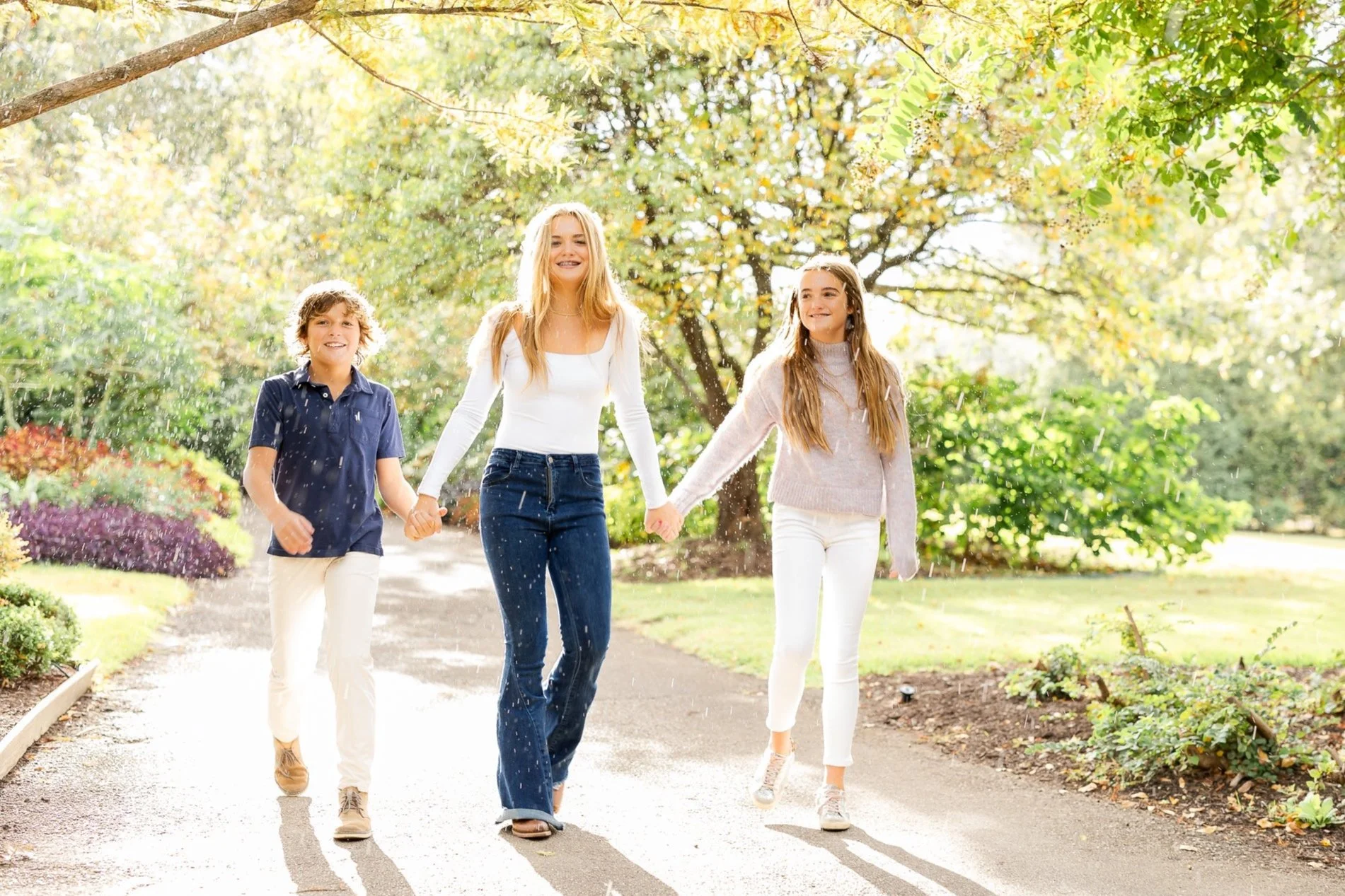 Three children walking hand in hand through a park on a sunny day, surrounded by trees and greenery.