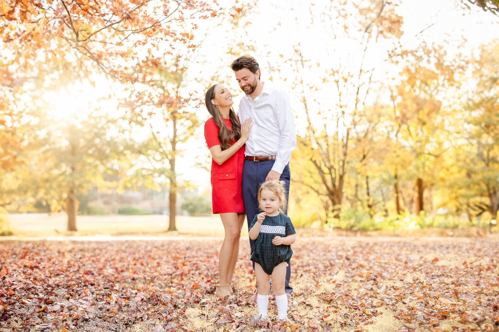 Family portrait in an autumn park, with a couple standing and a toddler in the foreground, surrounded by fallen leaves and trees with colorful foliage.