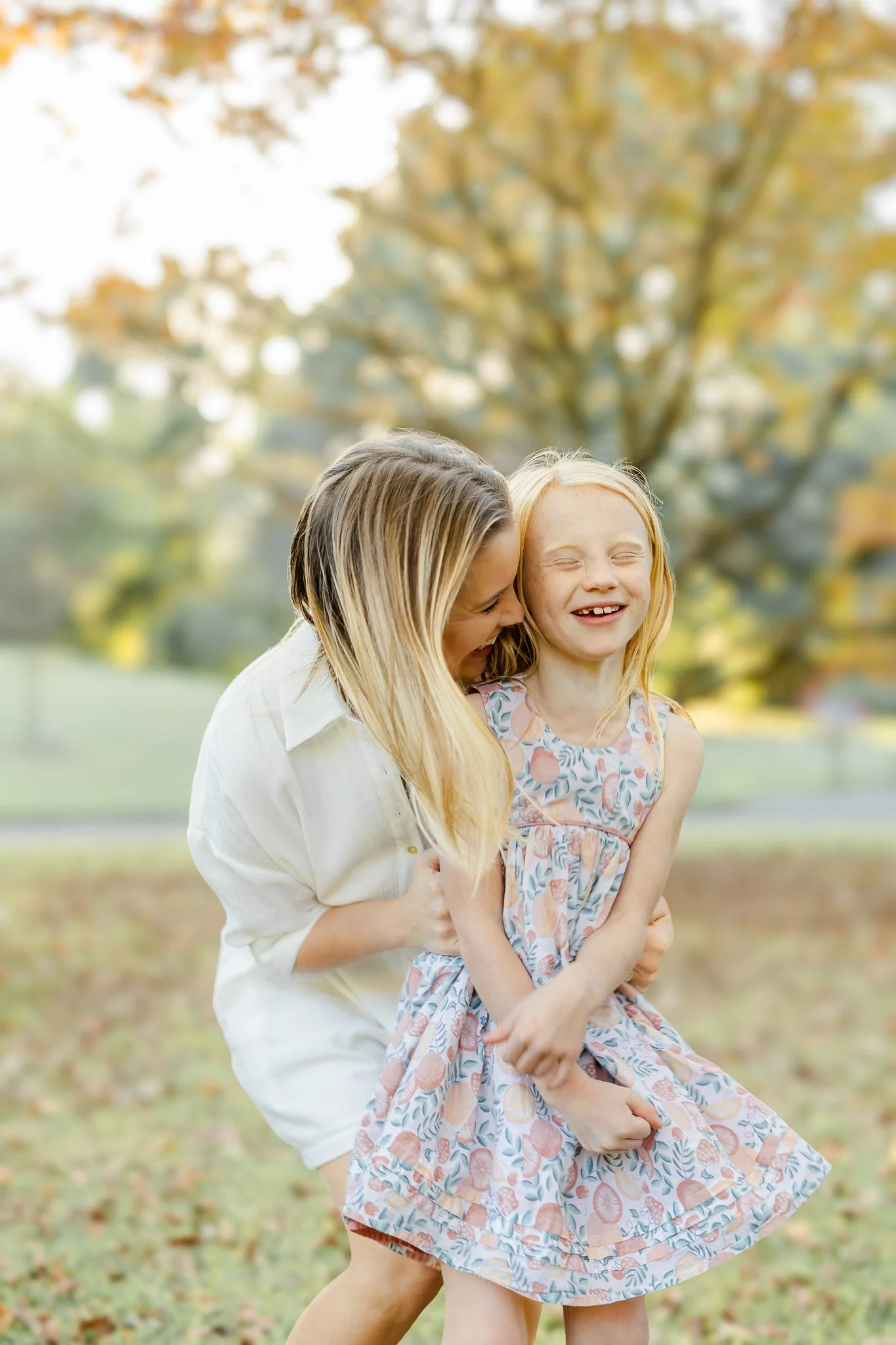 Two smiling children, an older girl hugging a younger girl, standing outdoors in a field during autumn. The younger girl is wearing a floral dress.