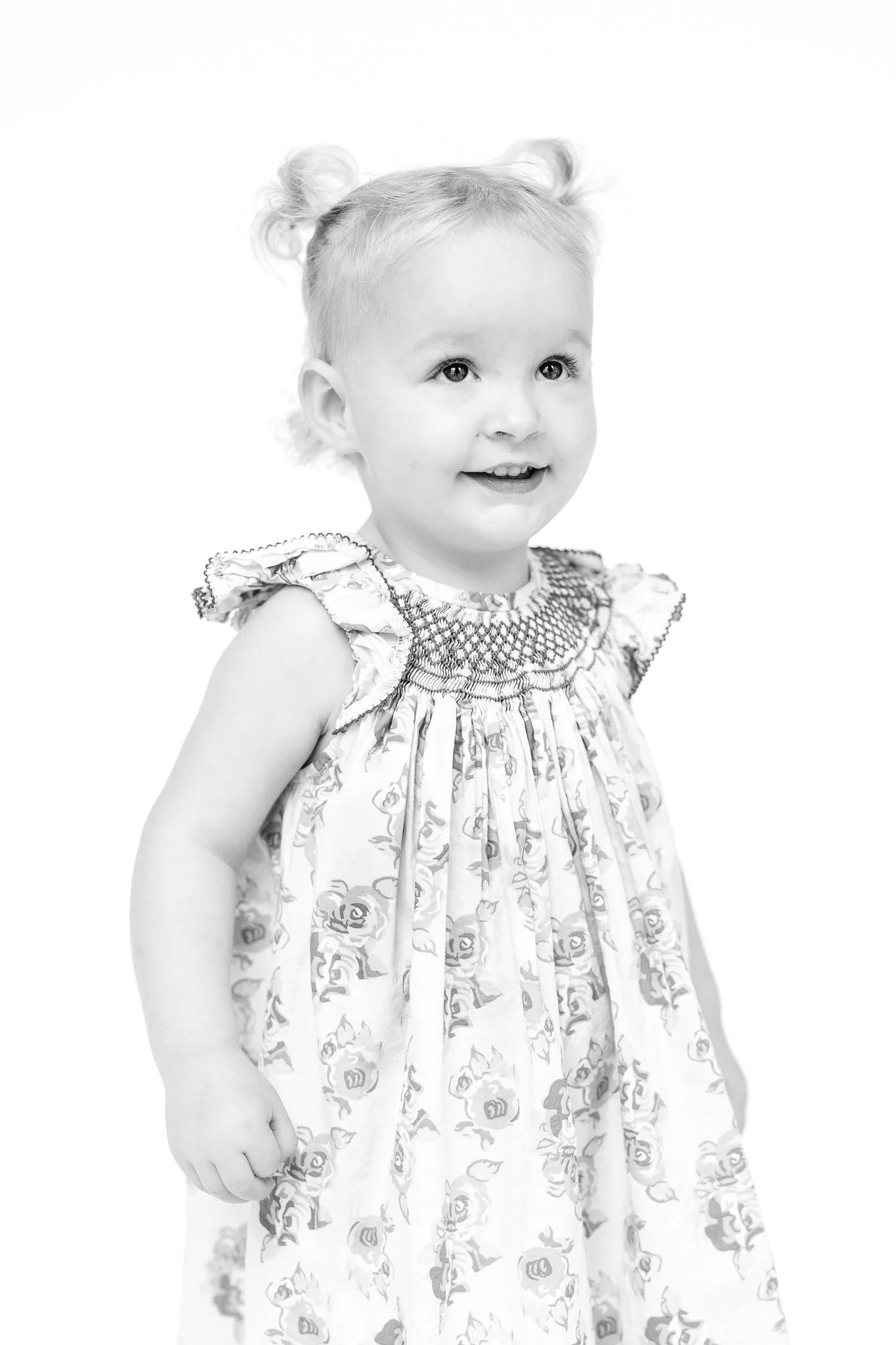 Black and white photo of a young child with light hair in pigtails wearing a patterned dress, standing and smiling.