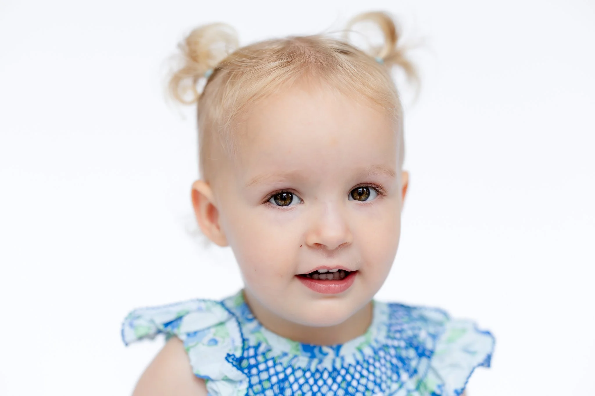 Portrait of a toddler with blonde hair and pigtails, wearing a blue patterned dress, smiling slightly against a white background.