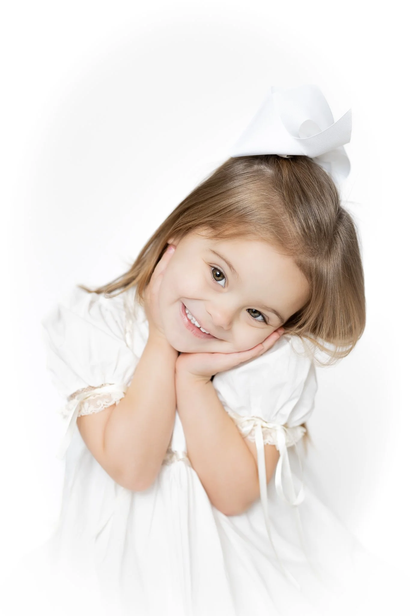 Smiling young girl with long hair, white dress, and large white bow posing with hands on cheeks.