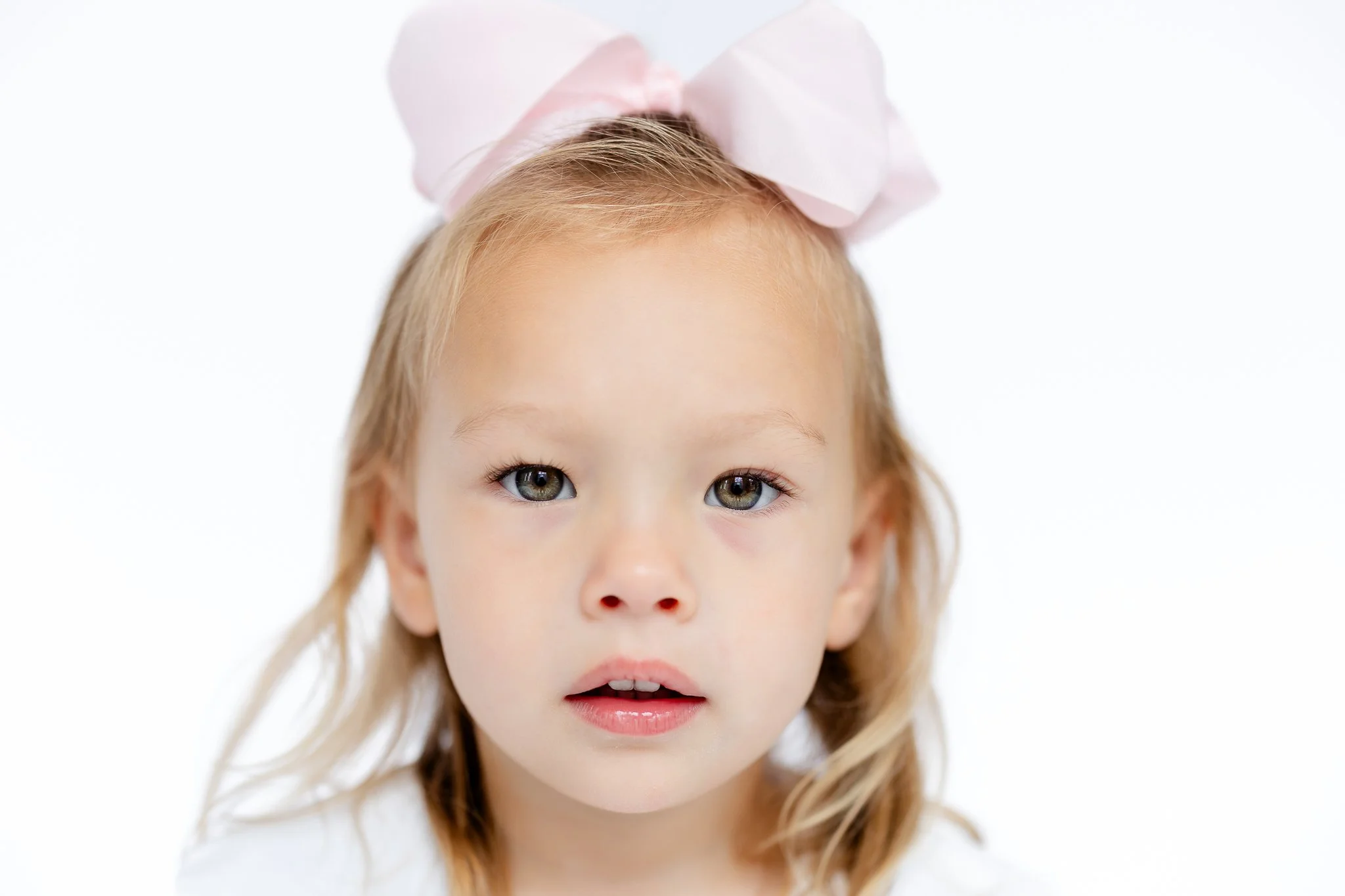 Young child with a pink bow in her hair, looking directly at the camera, against a white background.