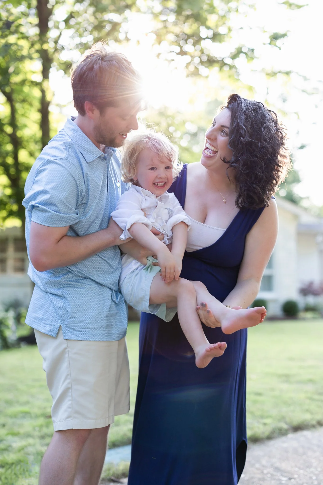 A family of three smiling outdoors with sunlight filtering through trees. The man is holding a child while the woman stands beside them. The setting is a grassy yard with trees and a house in the background.