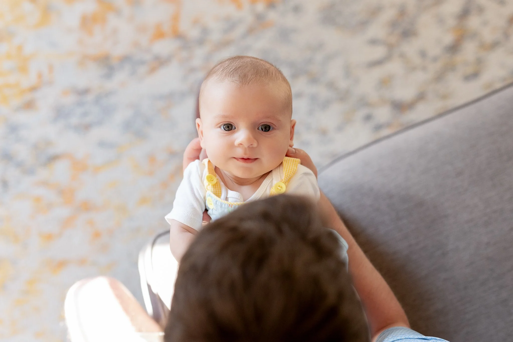 A baby wearing a white top and yellow overalls looking at a person with brown hair. The background is a carpet with a mix of colors.