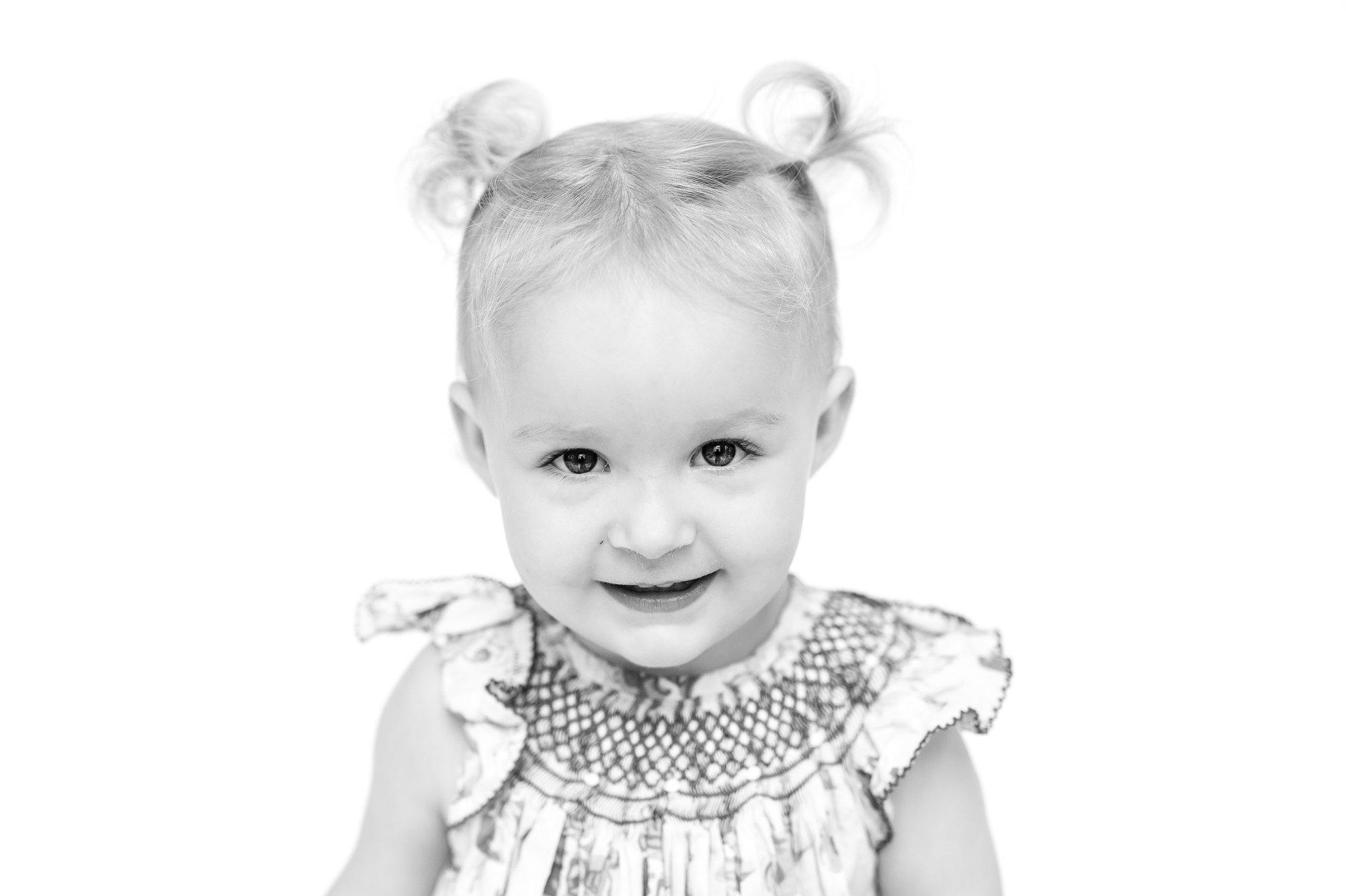 Black and white photo of a toddler with two small ponytails and a patterned dress, smiling at the camera.
