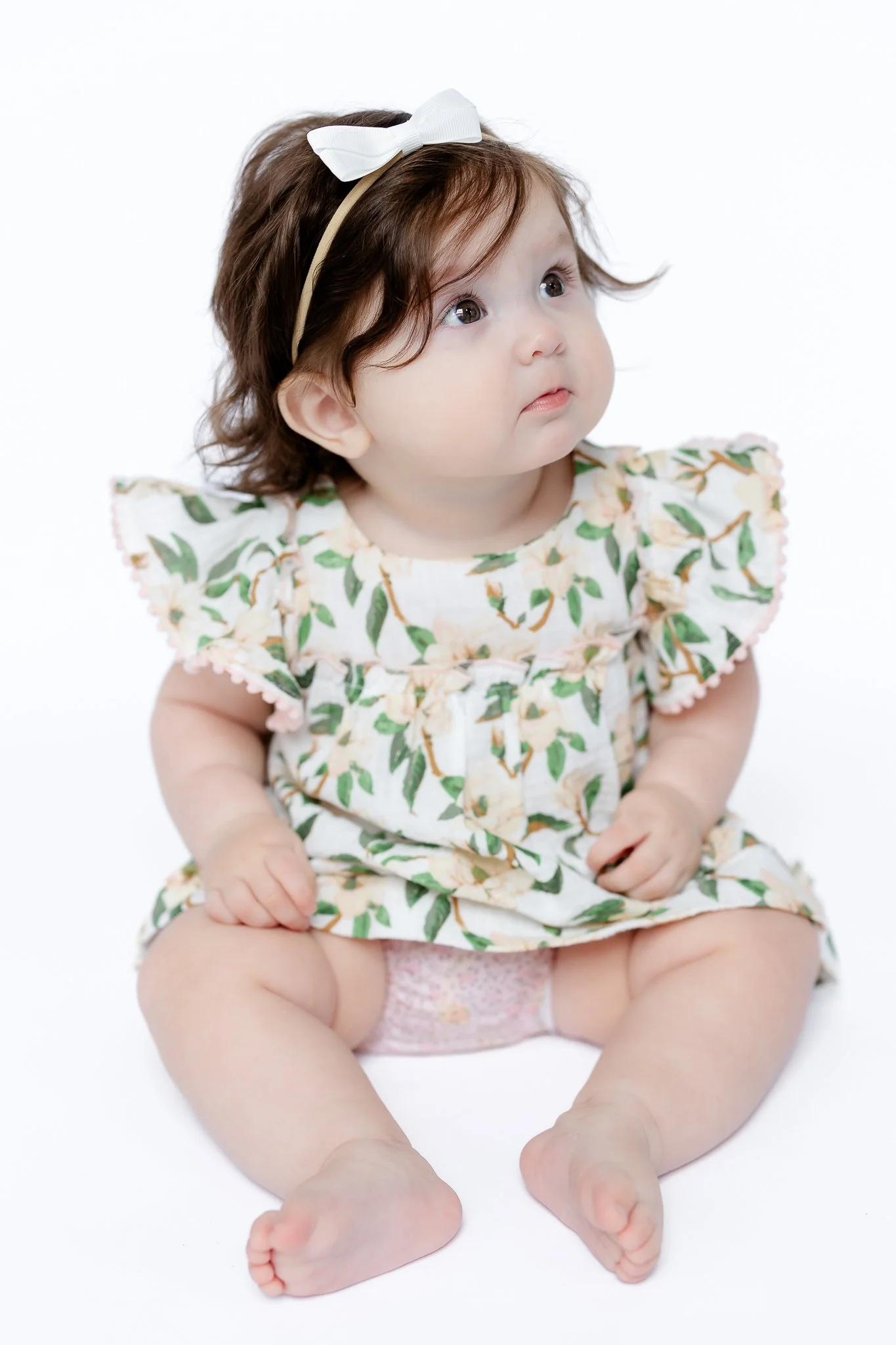 Baby sitting with a floral dress and white headband, looking upwards