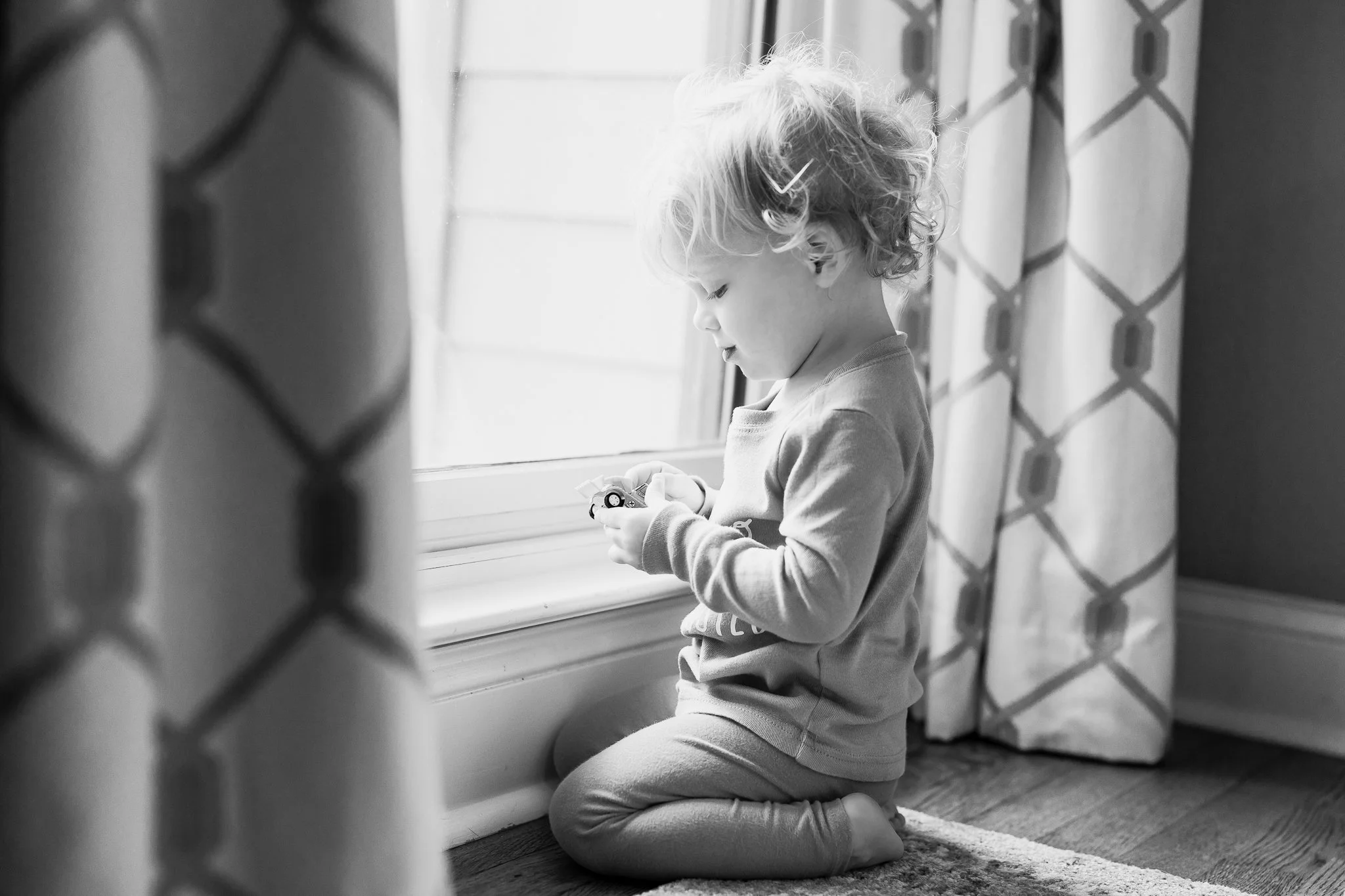 Black and white photo of a toddler in pajamas kneeling by a window playing with a toy car indoors.
