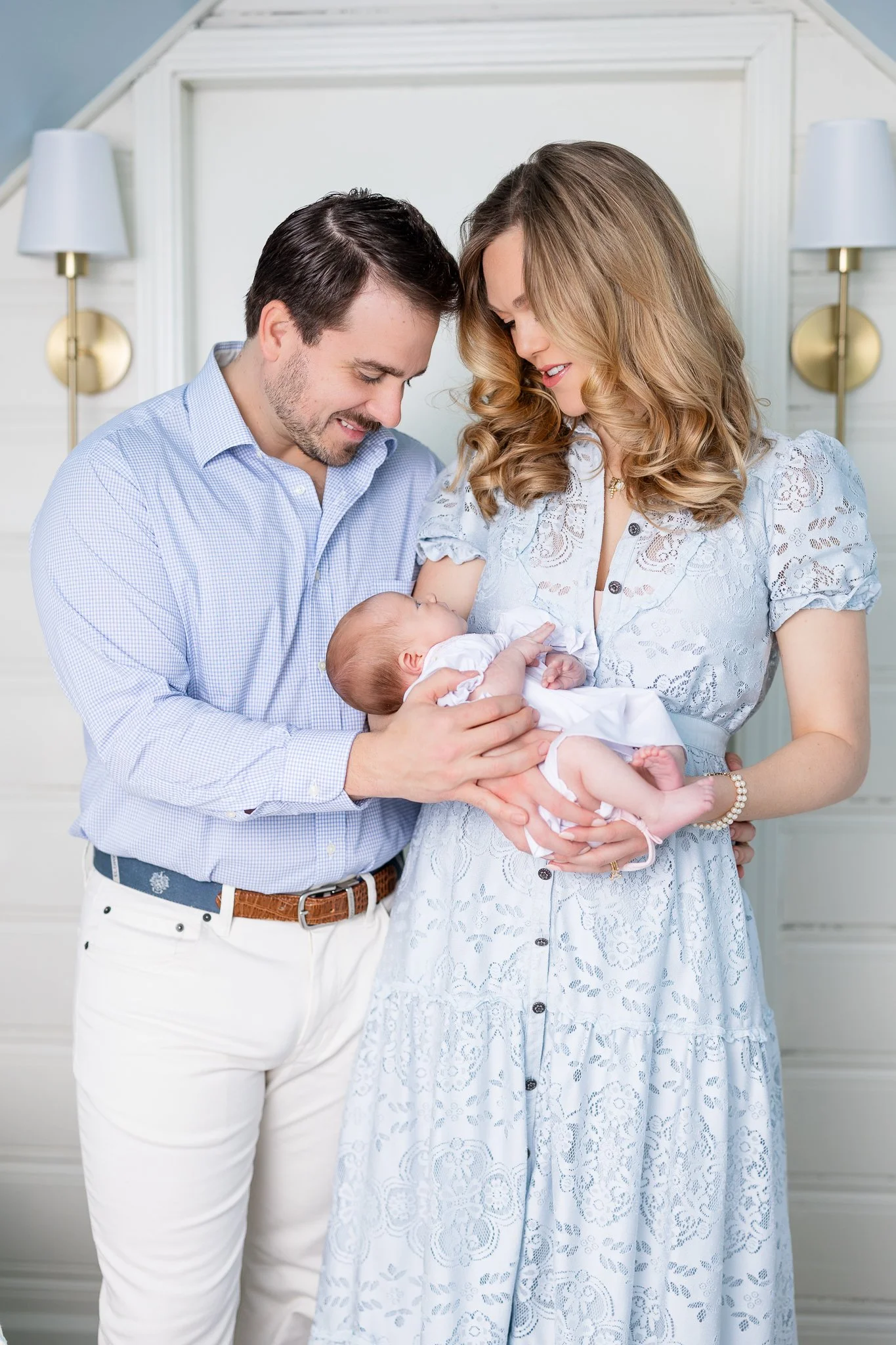 A couple dressed in light blue and white attire holding a baby, standing indoors with a soft, neutral background.