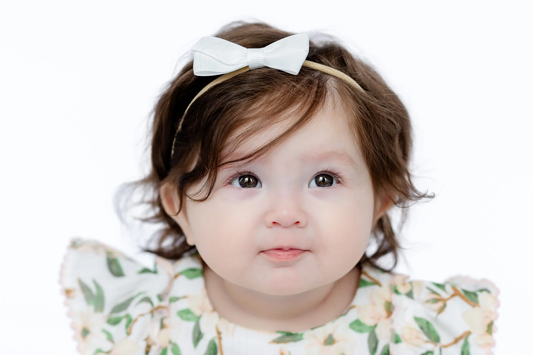 Close-up of a baby wearing a white headband with a bow, dressed in a floral outfit.