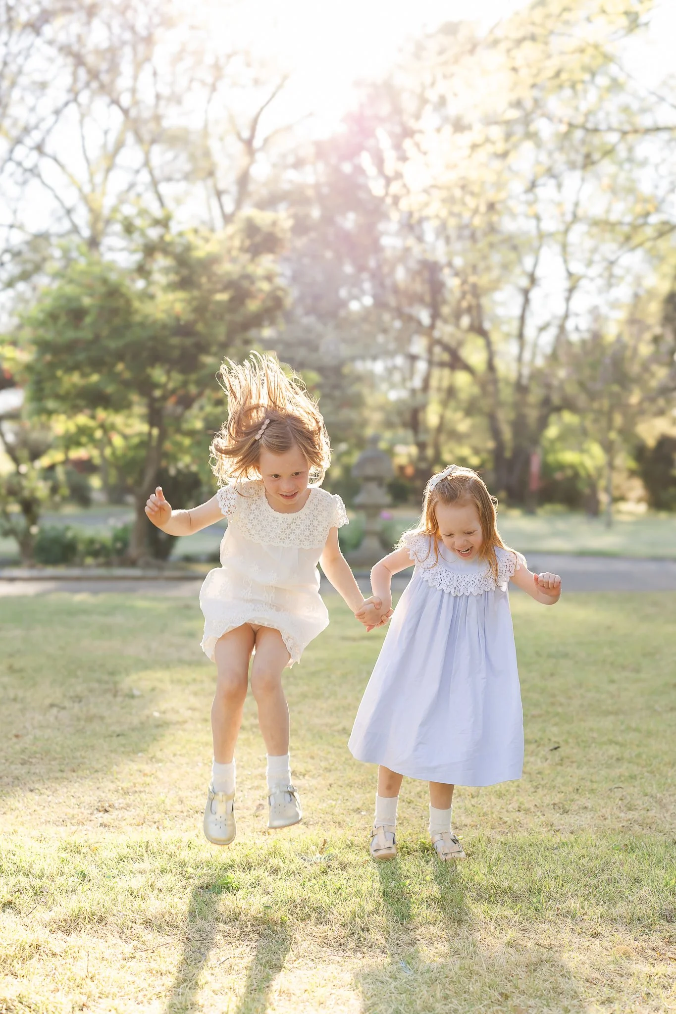Two young girls in white dresses playing and jumping on grass in a sunny park.