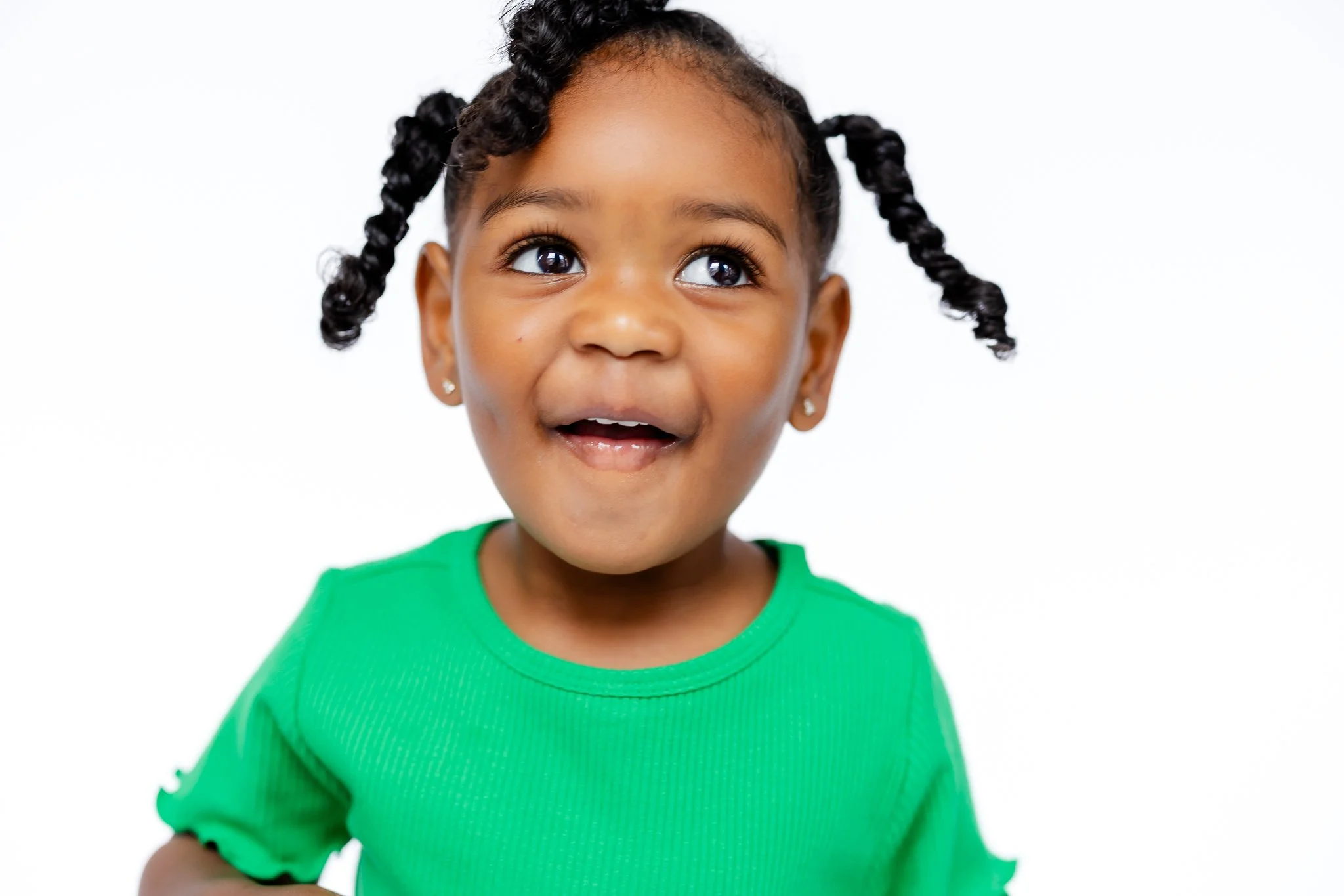 Smiling child with braided hair wearing a green shirt, looking upward against a white background.