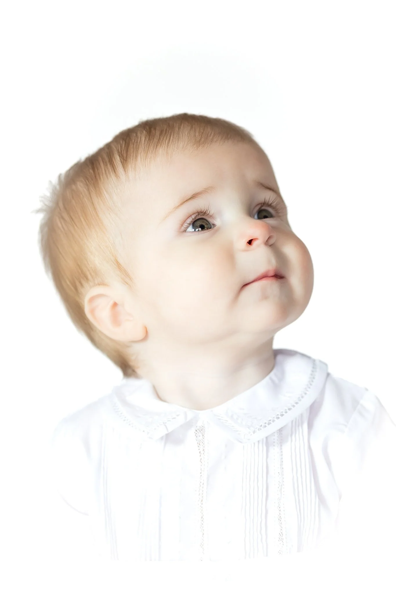 Close-up of a baby looking upwards, wearing a white collared shirt, against a white background.