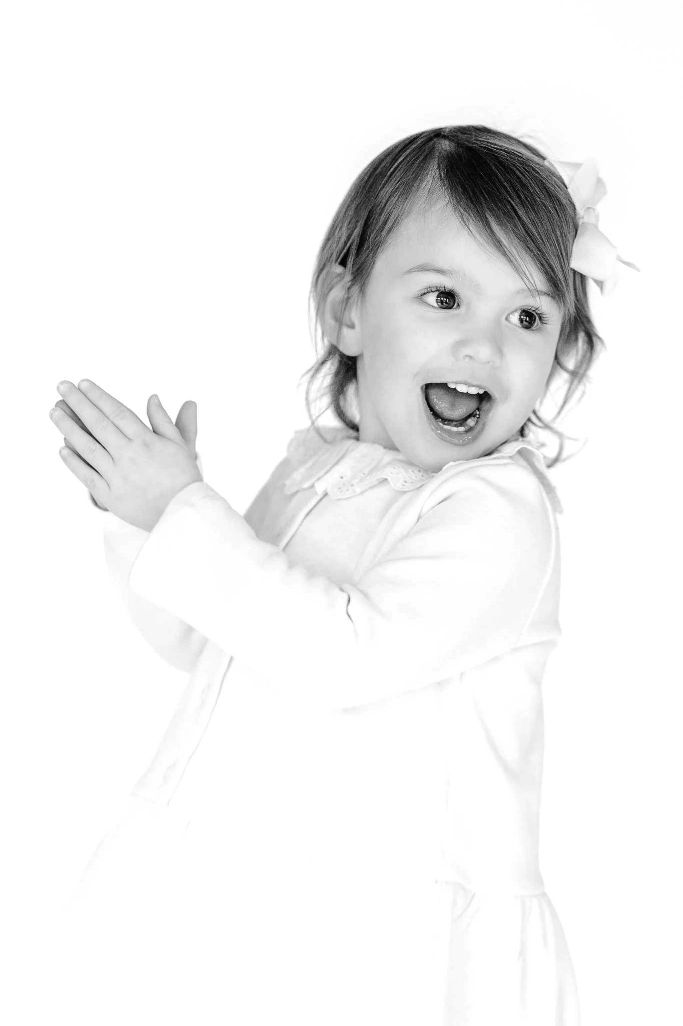Black and white photo of a young child smiling and clapping, wearing a white outfit and a bow in her hair.