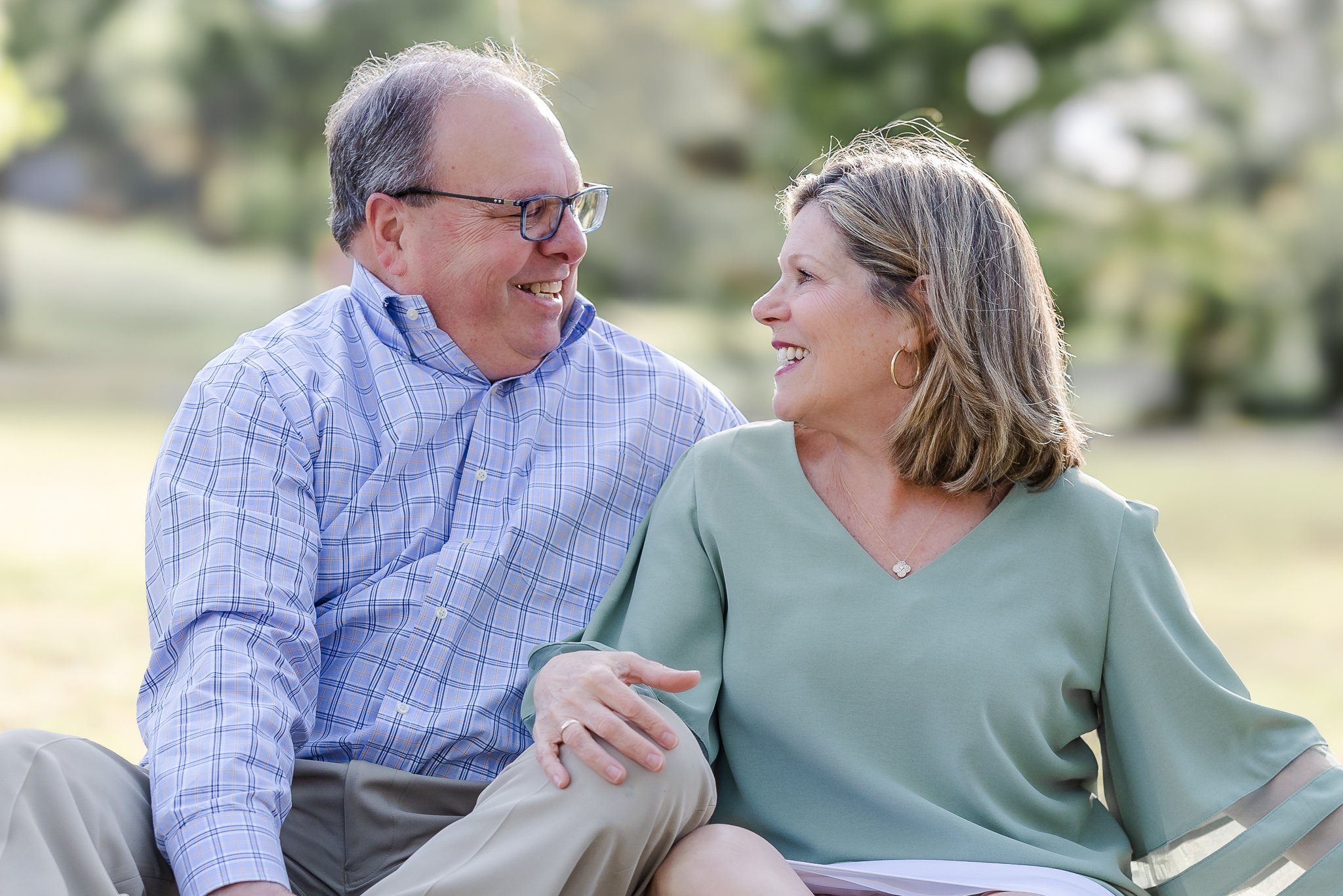A smiling man and woman sitting closely outdoors, enjoying each other's company. The man is wearing glasses and a blue checked shirt, and the woman is wearing a green top. They are both looking at each other happily.