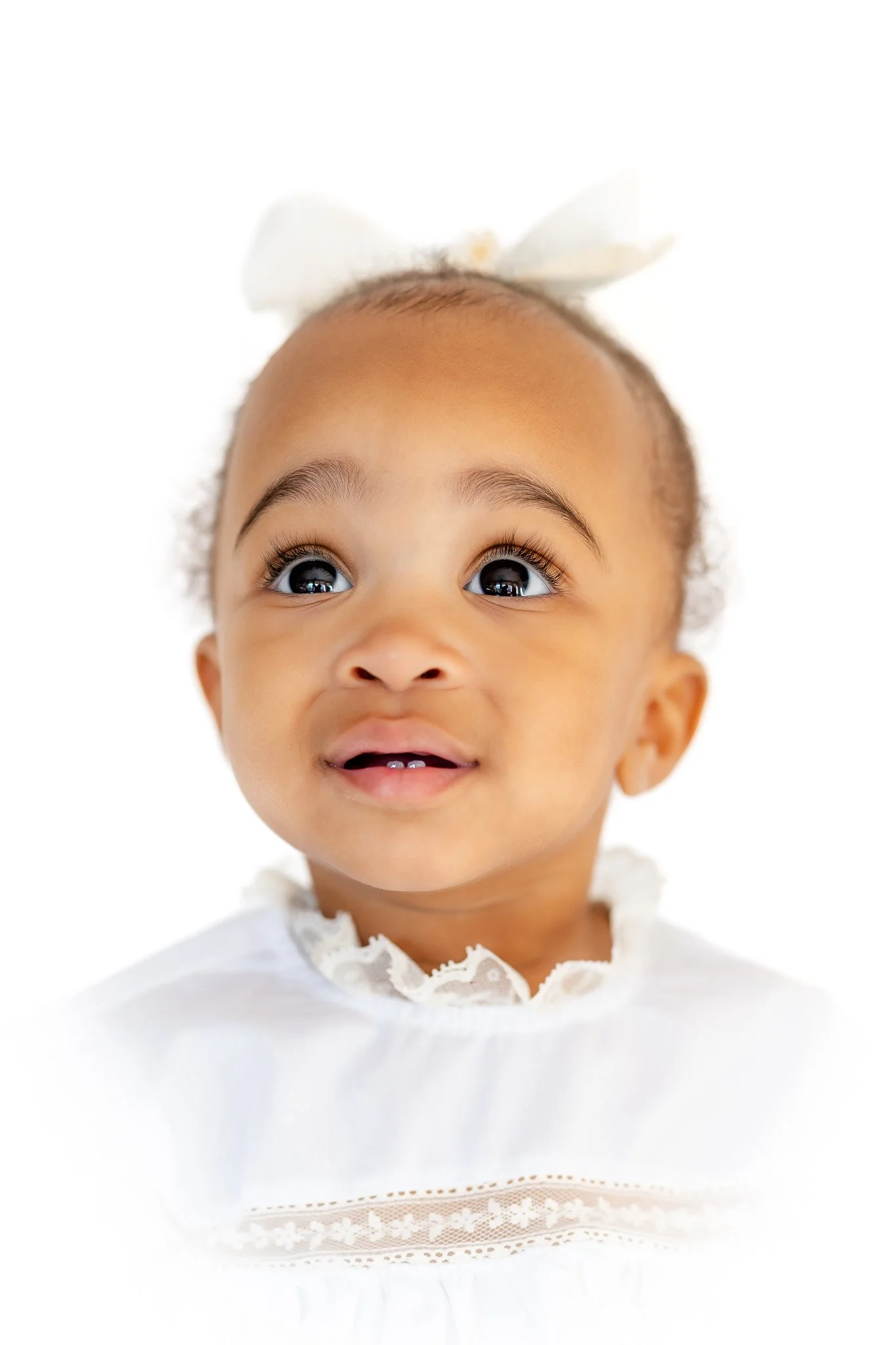 Portrait of a baby with a white bow and lace-trimmed outfit, looking upwards with a curious expression.