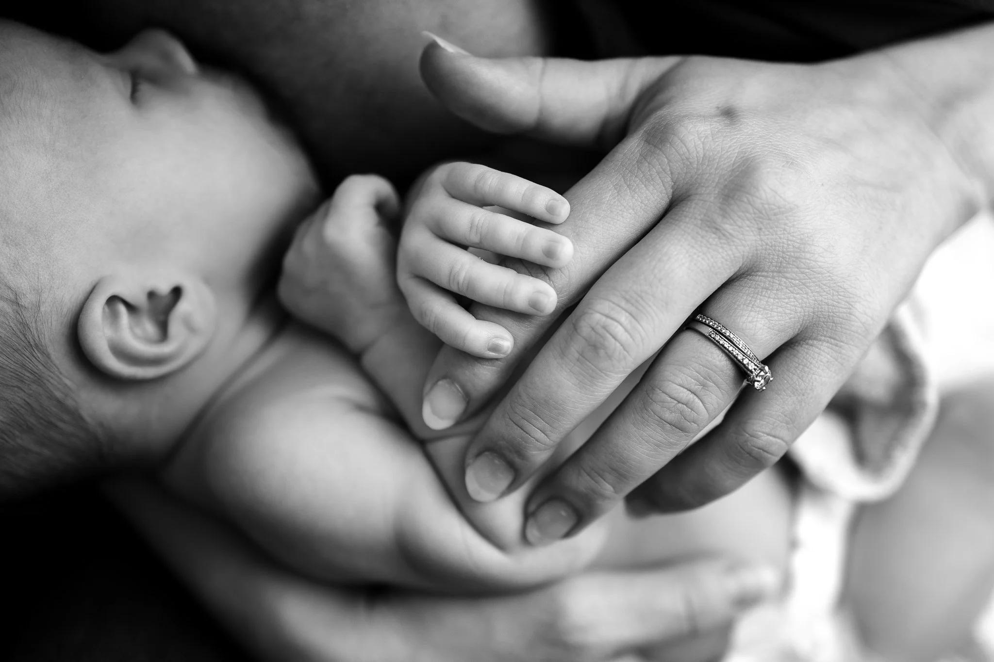 Black and white image of a baby sleeping, holding an adult's hand wearing a ring.