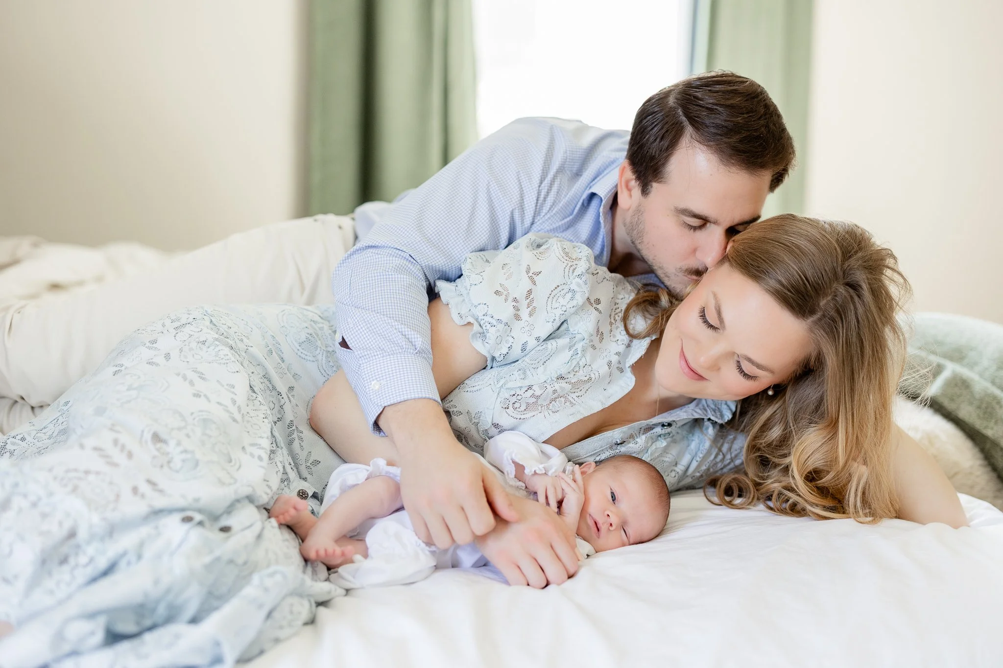 A couple lying on a bed with a newborn baby, the man kissing the woman's forehead.