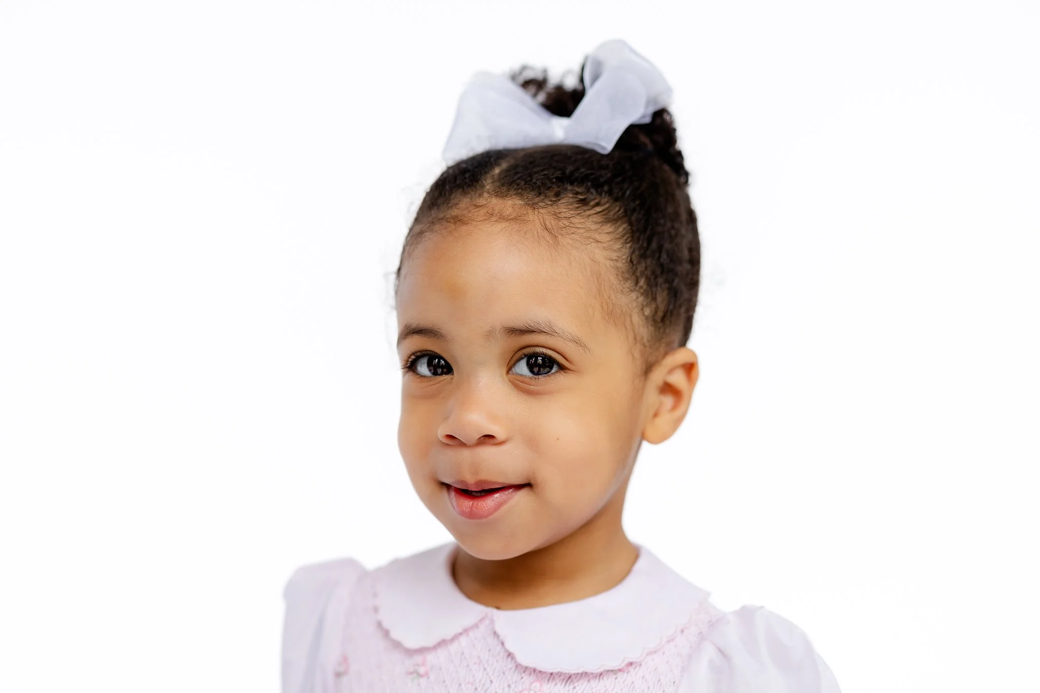 Smiling child with white bow and pink dress against white background.