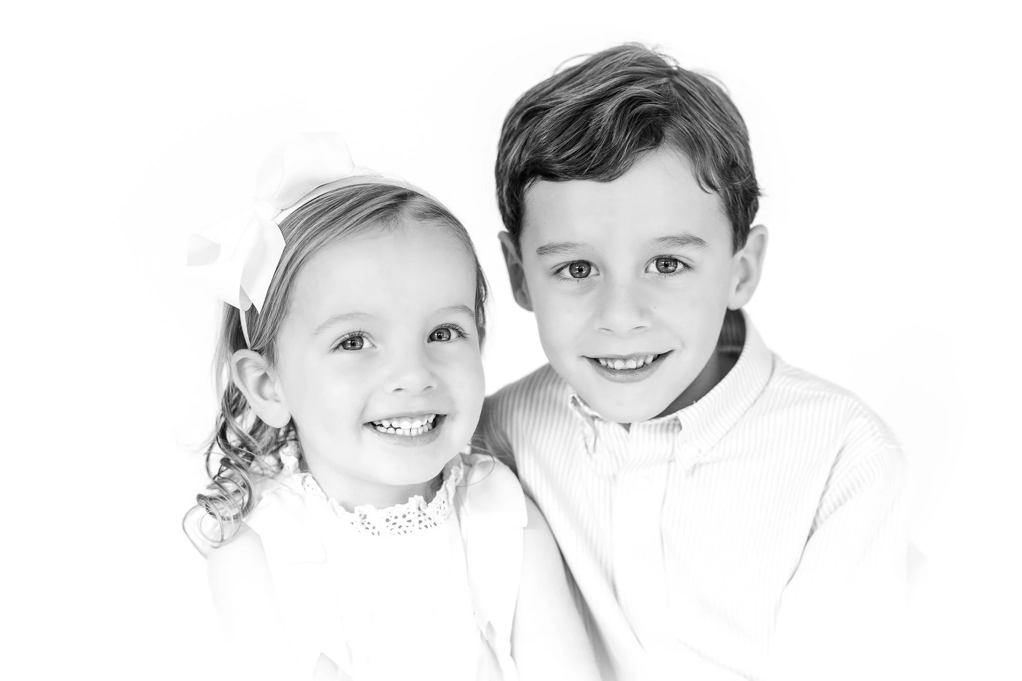 Black and white portrait of two smiling children, a young girl with a bow in her hair and an older boy wearing a striped shirt.
