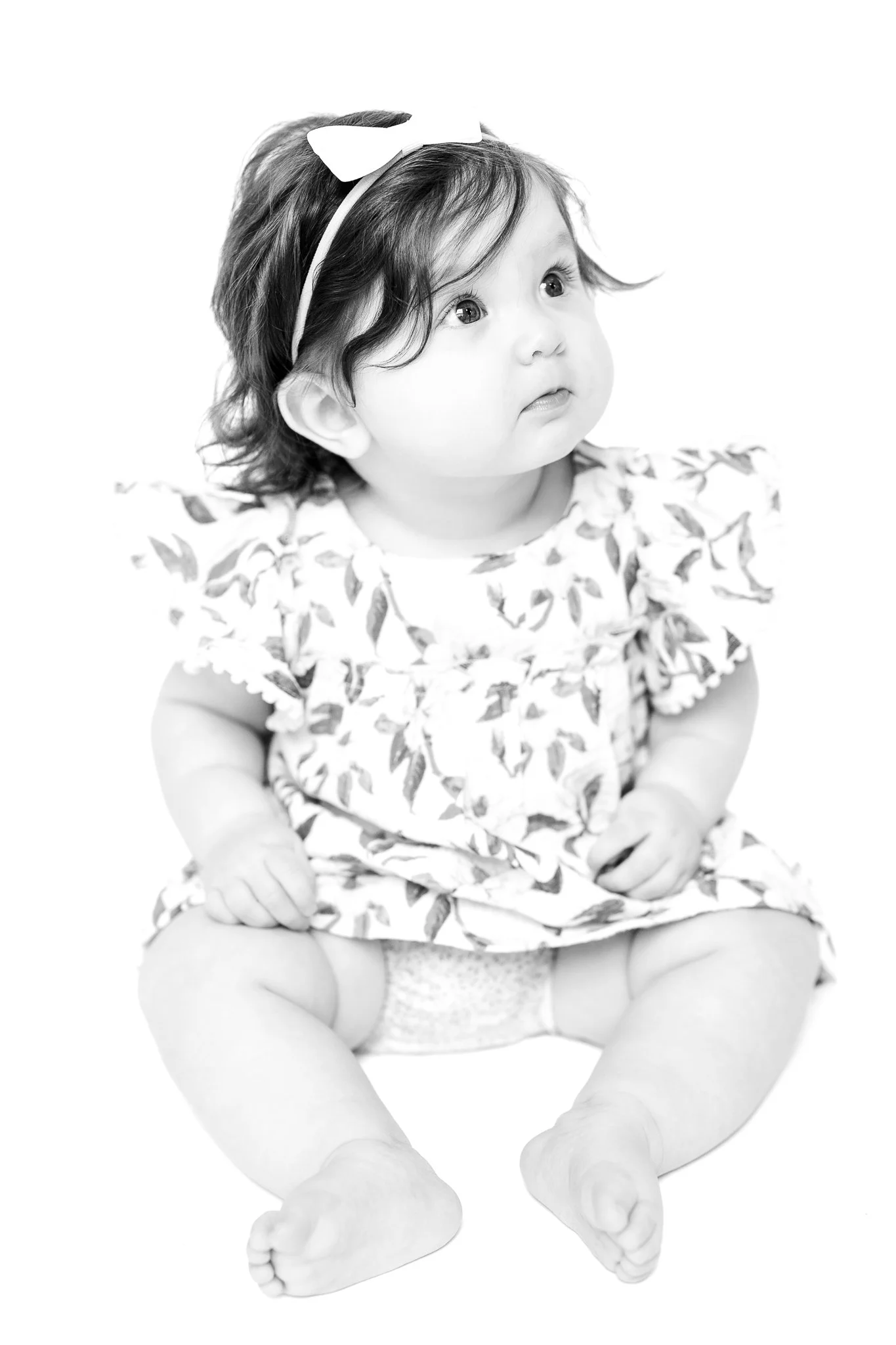 Black and white photo of a baby sitting, wearing a floral dress and a headband with a bow.