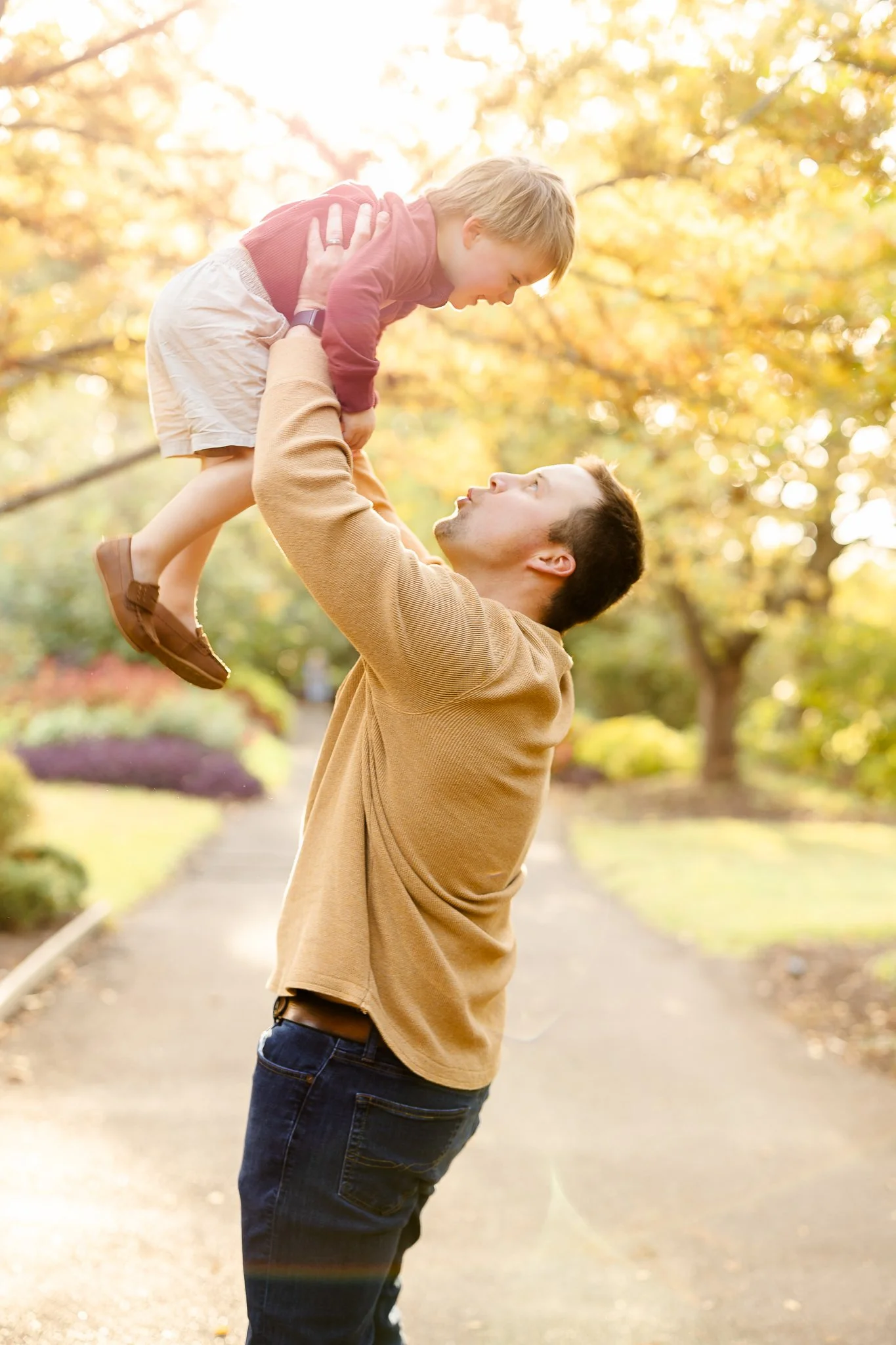 A man lifting a child in a park with autumn leaves.