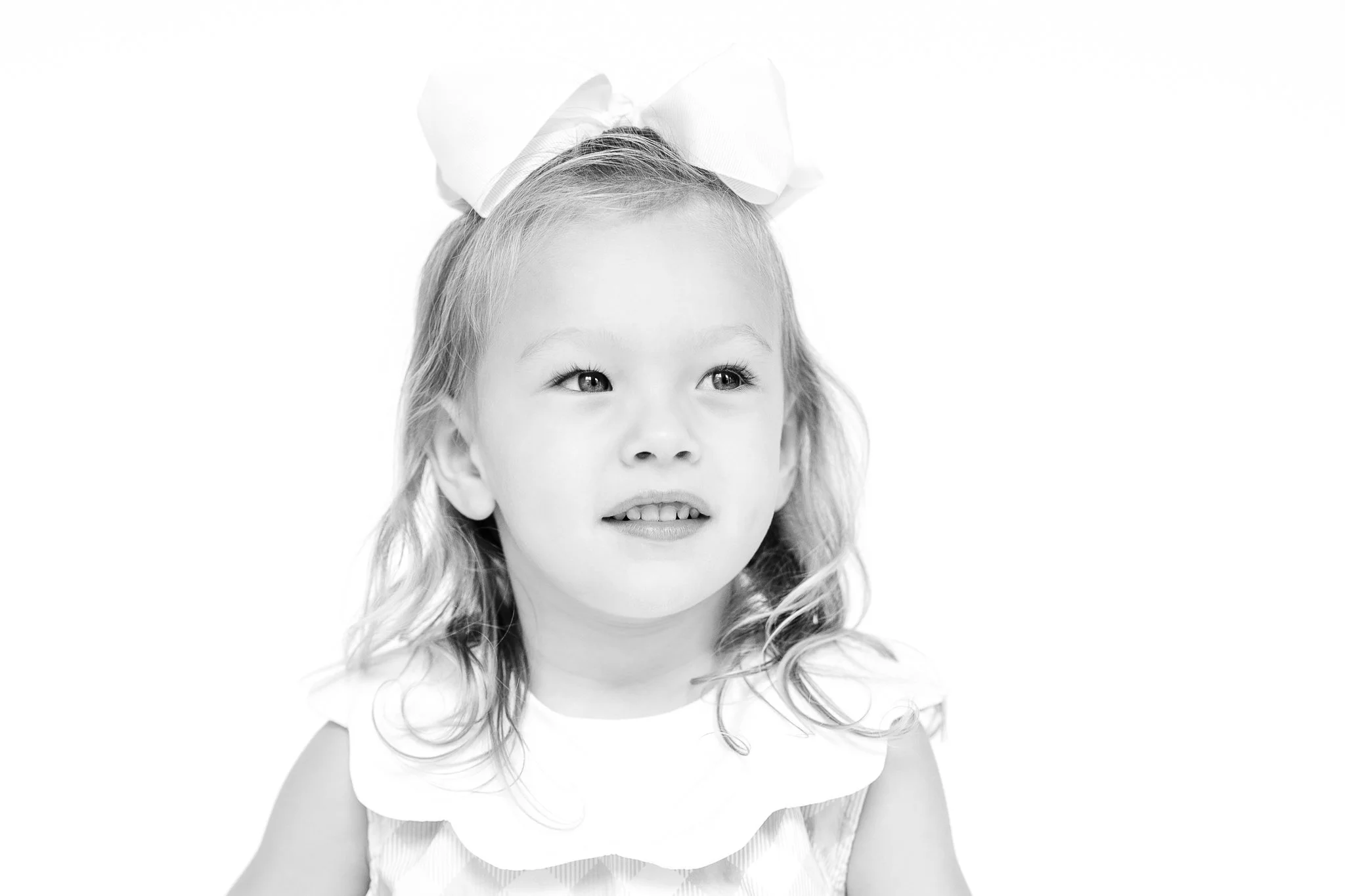 Black and white portrait of a young girl with a large bow in her hair, smiling and looking to the side.