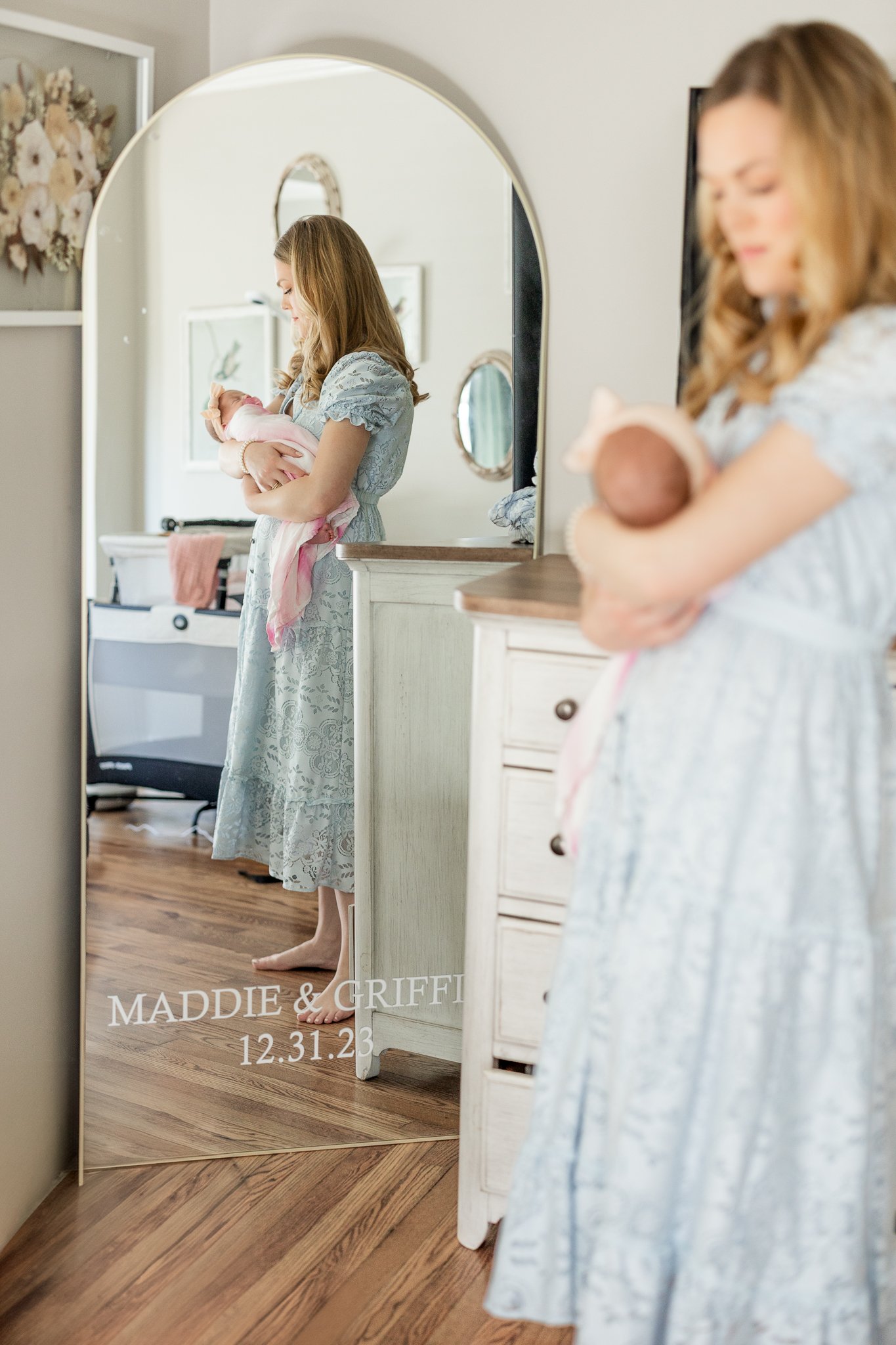 Woman in blue dress holding baby in a room with a large mirror and wooden floor, reflected in the mirror.
