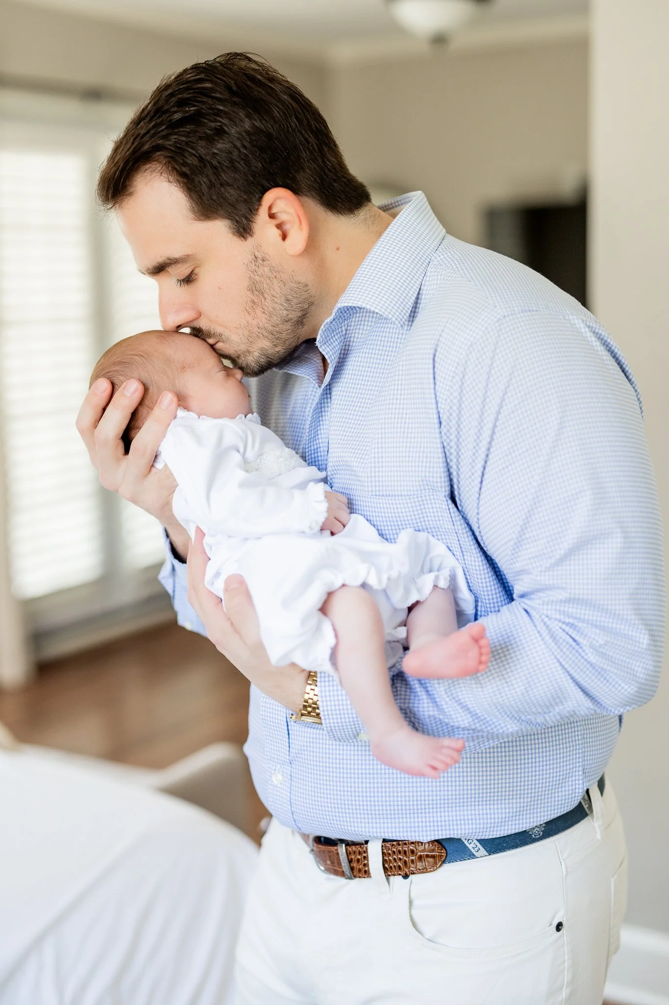 A man in a blue checkered shirt kisses a newborn baby, who is wearing white clothing, in a home setting.