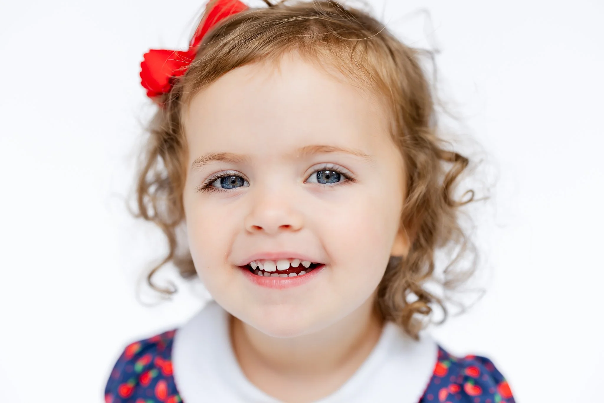 Smiling child with curly hair and a red bow, wearing a patterned dress, on a white background.