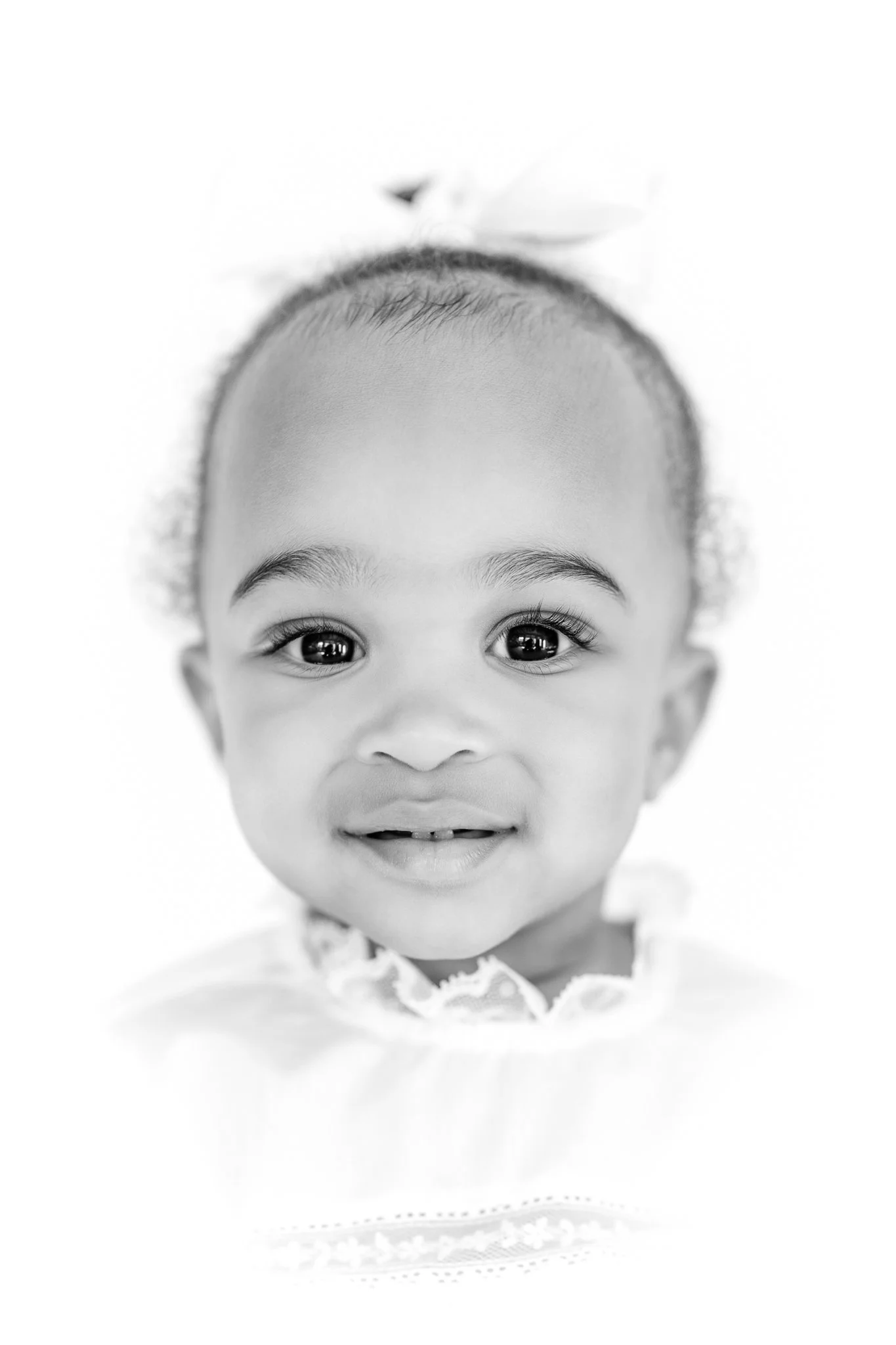 Black and white photo of a smiling toddler with curly hair and a bow.