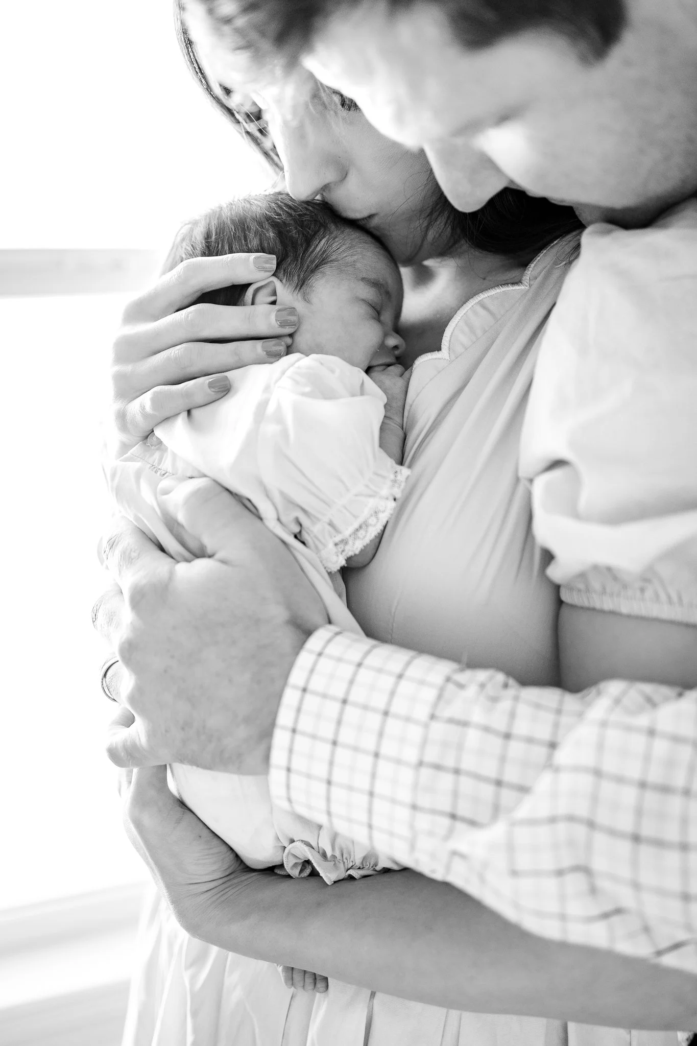 Black and white photo of parents holding a newborn baby, with the mother kissing the baby's head.